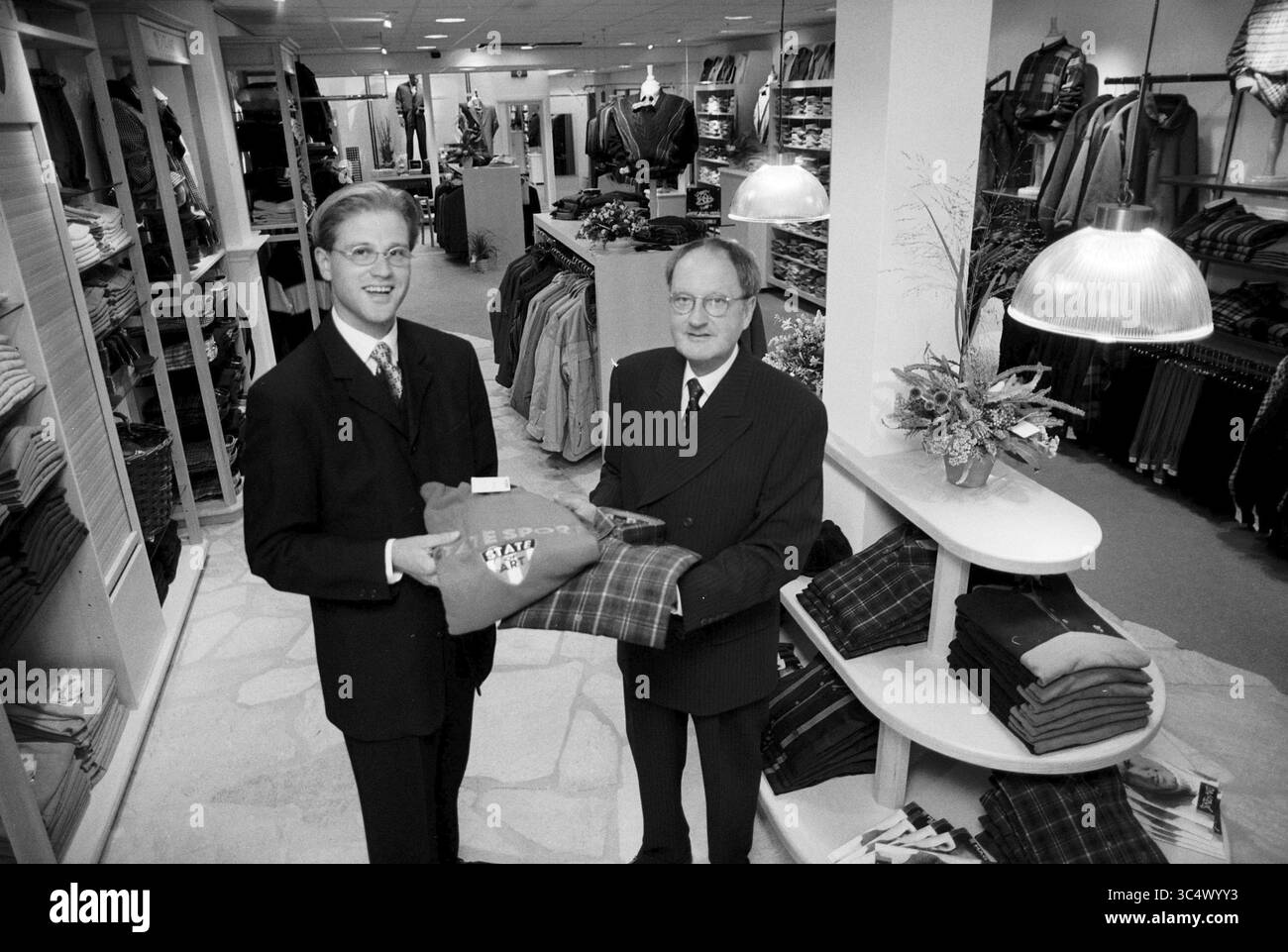 Van Keulen Mode, IJm., IJmuiden, Niederlande, 27-08-1996 Whizgle News, Dutch Desk, Niederlande, 1950-2000 zwei Männer in formeller Kleidung stehen in einem Bekleidungsgeschäft und lächeln, während einer dem anderen ein gefaltetes Hemd überreicht. Das Geschäft bietet ordentlich angeordnete Bekleidungsdisplays, die verschiedene Kleidungsstücke im Hintergrund präsentieren. Stockfoto