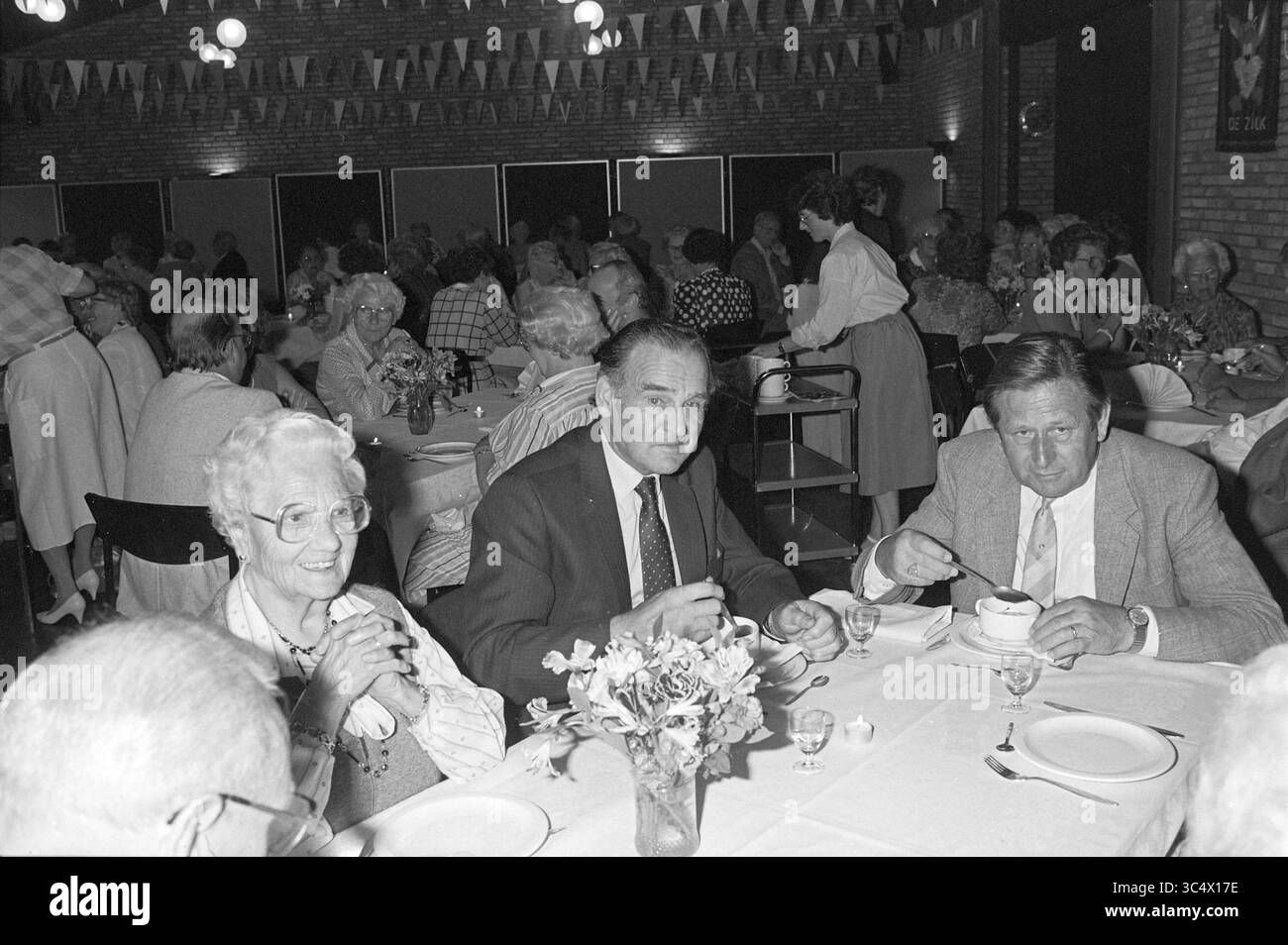 Seniors' Meal in the Zilk, Food and Eateries, 03-10-1985 Whizgle News, Dutch Desk, Niederlande, 1950-2000 Ein lebhaftes Treffen füllt einen Raum mit dekorativen Fahnen, in dem die Gäste an elegant gedeckten Tischen speisen können. Drei Personen an vorderster Front führen Gespräche, wobei eine Frau warm lächelt, während zwei Männer einen aufmerksamen Moment bei ihren Mahlzeiten teilen. Die Atmosphäre ist festlich, die Teilnehmer unterhalten sich und genießen die Gesellschaft der anderen. Stockfoto