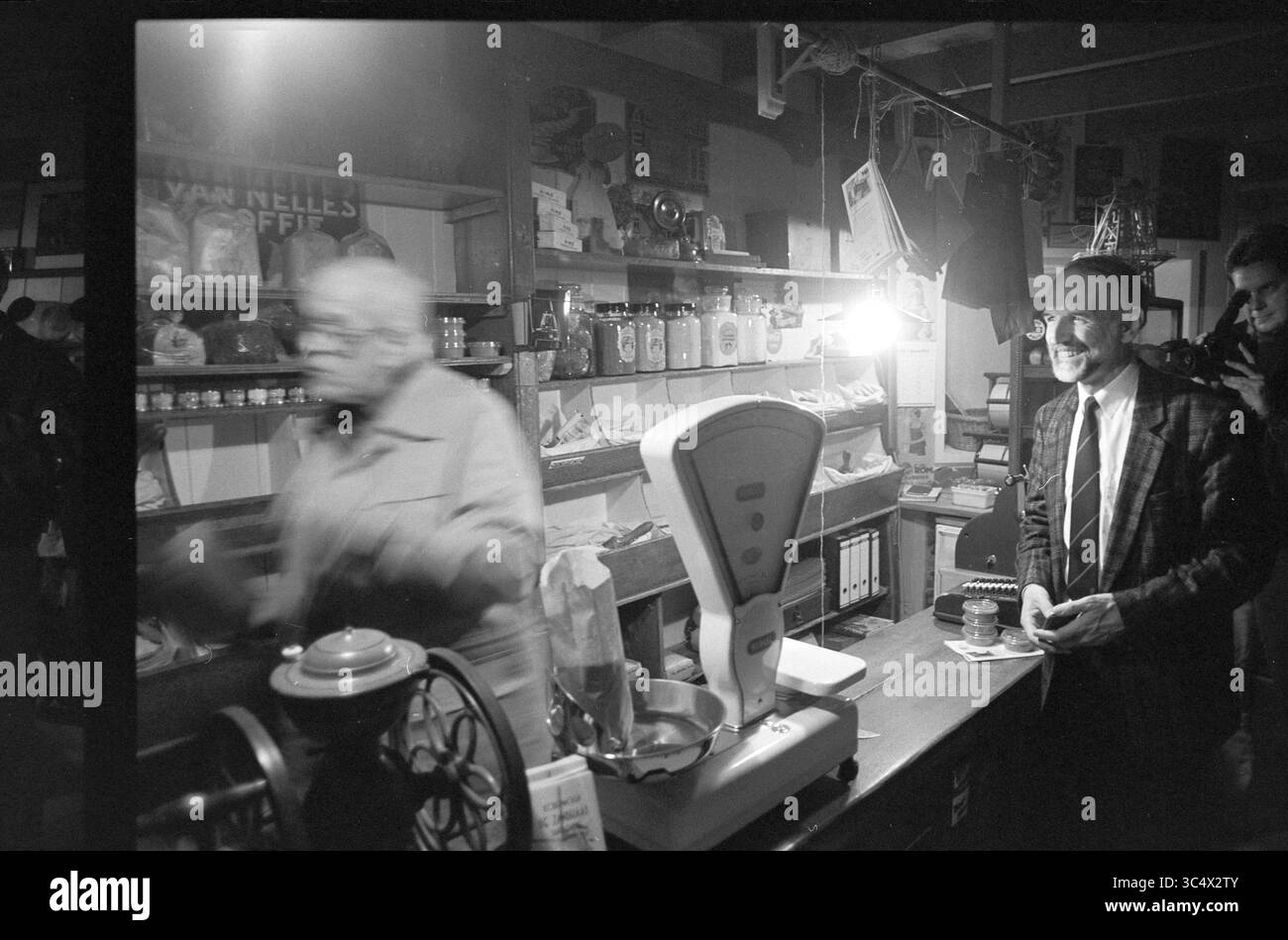Eröffnung Mehl Shop, S'poort, Santpoort, 05-11-1992 Whizgle News, Dutch Desk, Niederlande, 1950-2000 in einem hübschen Geschäft steht ein Mann in der Nähe einer Waage lächelnd, während ein anderer Kunde schnell vorbeizieht und ein Gefühl von geschäftiger Aktivität schafft. Regale mit Gläsern und verpackten Waren spiegeln einen nostalgischen Charme wider und deuten auf eine vergangene Ära des lokalen Handels hin. Stockfoto