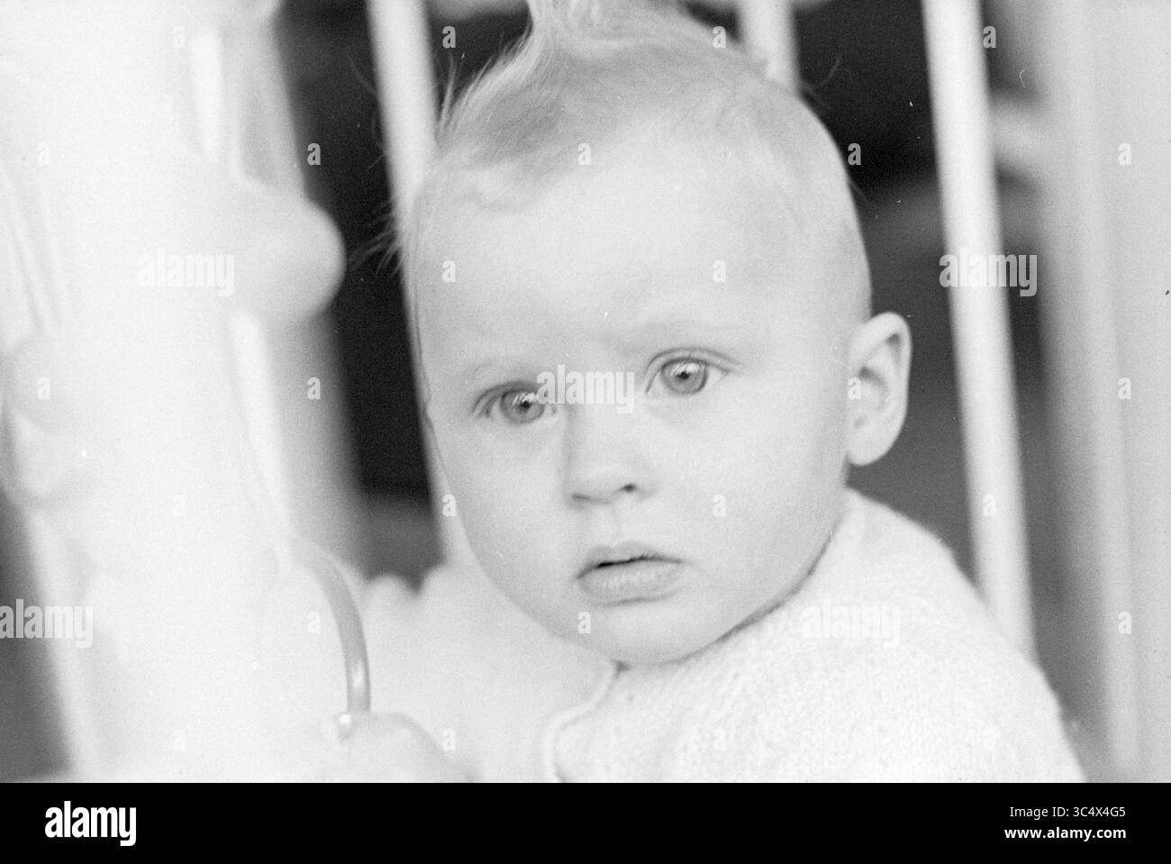 Baby in Playpen Whizgle News, Dutch Desk, Niederlande, 1950-2000 Ein neugieriges Kind blickt mit hellen Augen und einer unverwechselbaren Frisur, während es sich an einer verspielten Struktur in der Nähe festhält. Stockfoto