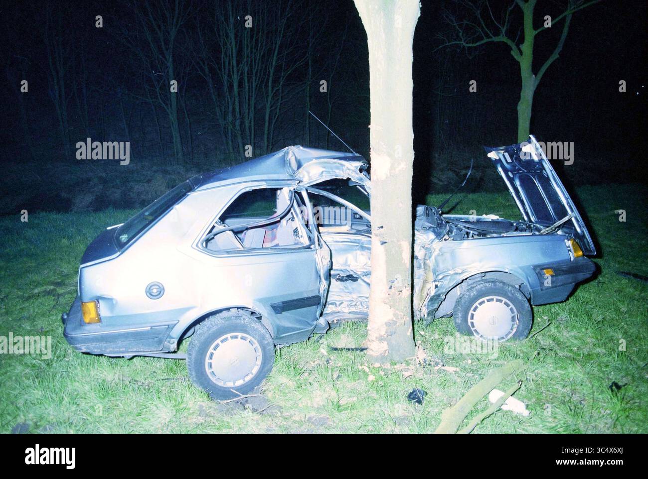 Tödlicher Unfall Amsterdamseweg IJmuiden, Amsterdam, Amsterdam, Amsterdamseweg, Niederlande, 11-03-1998 Whizgle News, Dutch Desk, Niederlande, 1950-2000 Ein beschädigtes Auto wird an einem Baum festgeklemmt und zeigt die Auswirkungen einer Kollision. Der zerknitterte Körper und die offene Haube zeigen das Ausmaß der Zerstörung, während die Umgebung schwach beleuchtet ist, was die düstere Atmosphäre der Szene noch verstärkt. Stockfoto