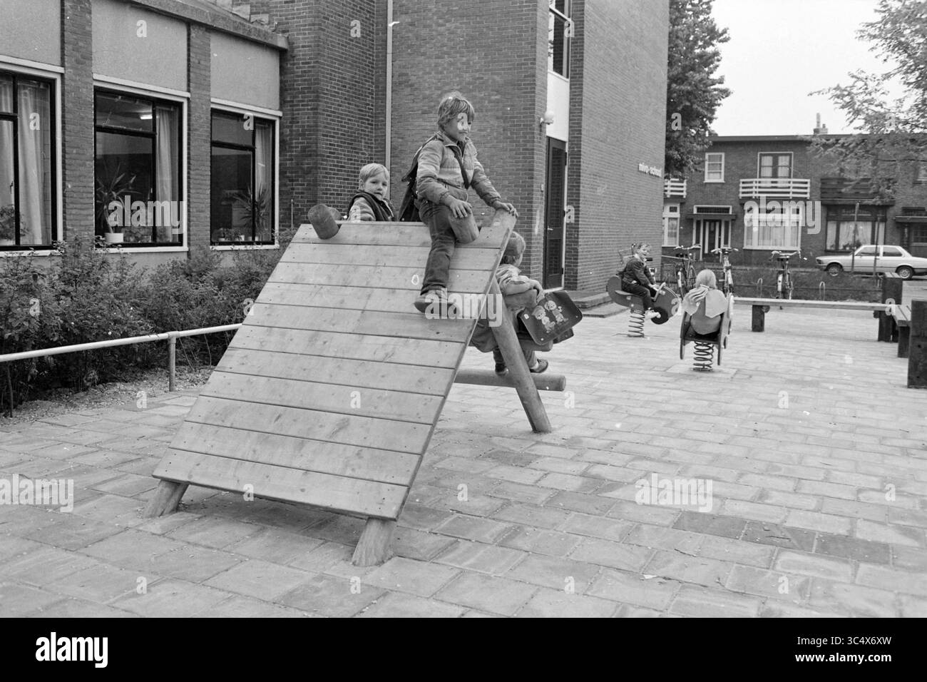 Spielgeräte in der MIVO School, IJmuiden, Spiele und Spielzeug, Schulen, IJmuiden, Niederlande, 25-05-1984 Whizgle News, Dutch Desk, Niederlande, 1950-2000 Eine Gruppe von Kindern spielt auf einem Schulhof, wobei ein Kind eine Holzrutsche hinuntergleitet, während andere eine Federschaukel in der Nähe genießen. Die umliegenden Gebäude bieten eine Kulisse für einen erlebnisreichen Tag. Stockfoto