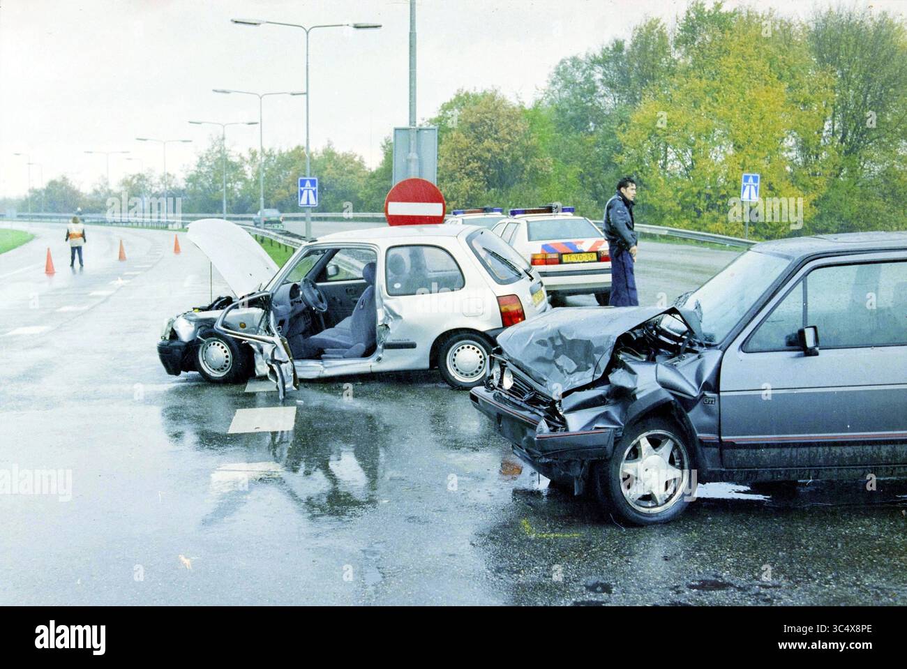 Unfall an der Ausfahrt Nieuwe Meer, 15-11-1995 Whizgle News, Dutch Desk, Niederlande, 1950-2000 zwei Autos sind in einen schweren Unfall auf einer nassen Straße verwickelt. Ein Fahrzeug weist erhebliche Schäden an der Vorderseite auf, während das andere mit angehobener Motorhaube ausgestattet ist. Das Notfallpersonal ist anwesend, verwaltet den Tatort und sorgt für Sicherheit, während die ausgestellten Verkehrskegel und Polizeifahrzeuge das Gebiet umgeben. Bäume im Hintergrund deuten auf eine vorstädtische Umgebung hin. Stockfoto