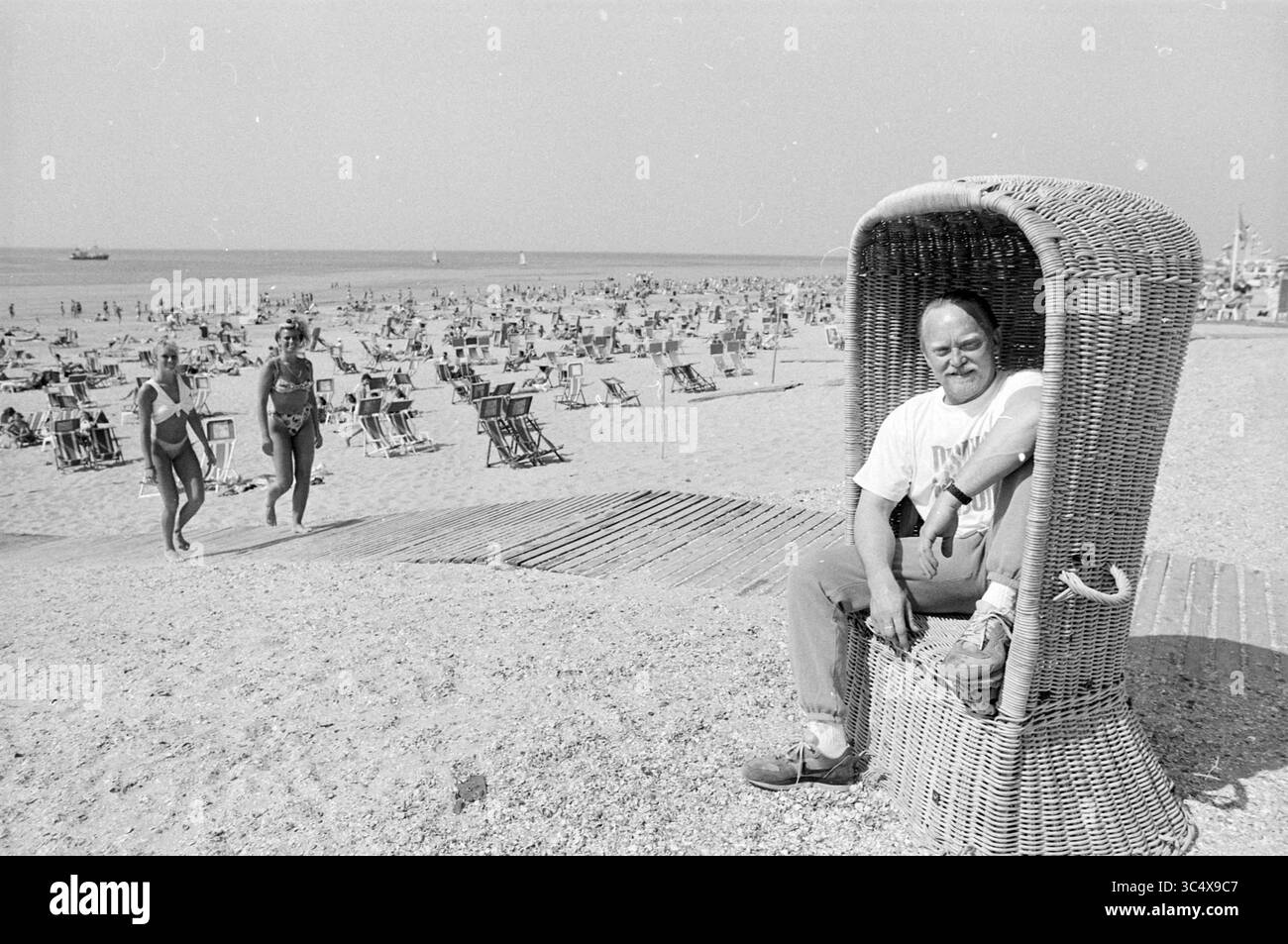 Verschiedene Aufnahmen Pavillon Tropical Zandvoort, Strand und Strände, Zandvoort, Zandvoort, 24-05-1989 Whizgle News, Dutch Desk, Niederlande, 1950-2000 Ein Mann sitzt bequem in einem großen Korbstuhl an einem belebten Strand, umgeben von Sonnenanbetern und Strandgängern, die den Sommertag genießen. Zwei Frauen schlendern vorbei und halten die entspannte Atmosphäre eines Kurzurlaubs am Meer fest. Stockfoto