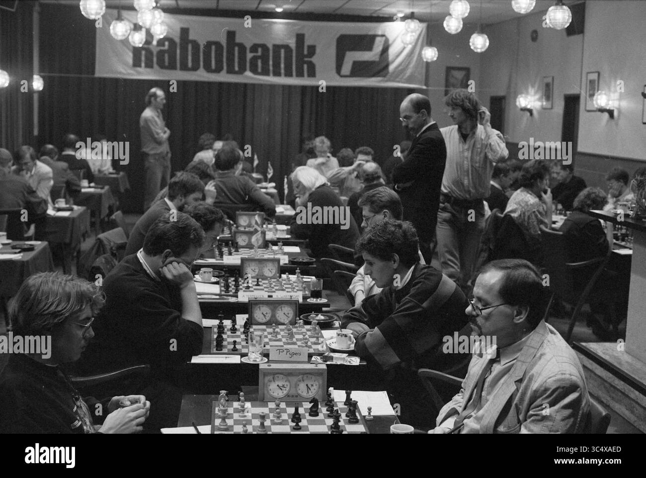 Schach in Zomerzorg, Hillegom, 31-10-1991, Whizgle News, Dutch Desk, Niederlande, 1950-2000 Ein hektisches Schachturnier in einer überfüllten Halle mit mehreren Schachbrettern für intensive Spiele. Die Teilnehmer sind sehr konzentriert und zeigen eine Mischung aus Konzentration und Wettkampfgeist, während die Zuschauer das Gameplay in einer festlichen Atmosphäre mit hängenden Laternen beobachten. Ein Banner im Hintergrund weist auf Sponsoring hin, was die Bedeutung der Veranstaltung erhöht. Stockfoto