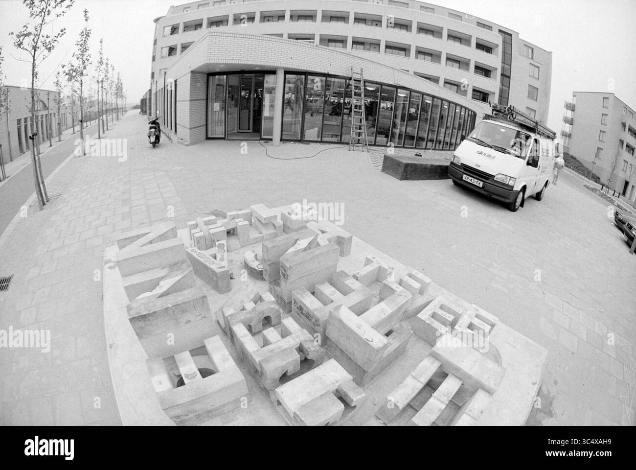 Abgerissene Briefe von Skulpturen vor der Bibliothek, Velserbroek, 01-06-1994 Whizgle News, Dutch Desk, Niederlande, 1950-2000 Eine große, abstrakte Skulptur, die einem fragmentierten Stadtbild ähnelt, befindet sich im Vordergrund, umgeben von neu errichteten Gebäuden und einem in der Nähe geparkten Van. Die Szene spiegelt ein städtisches Entwicklungsgebiet wider, mit Bäumen, die den Weg säumen, und Bautätigkeit im Hintergrund. Stockfoto