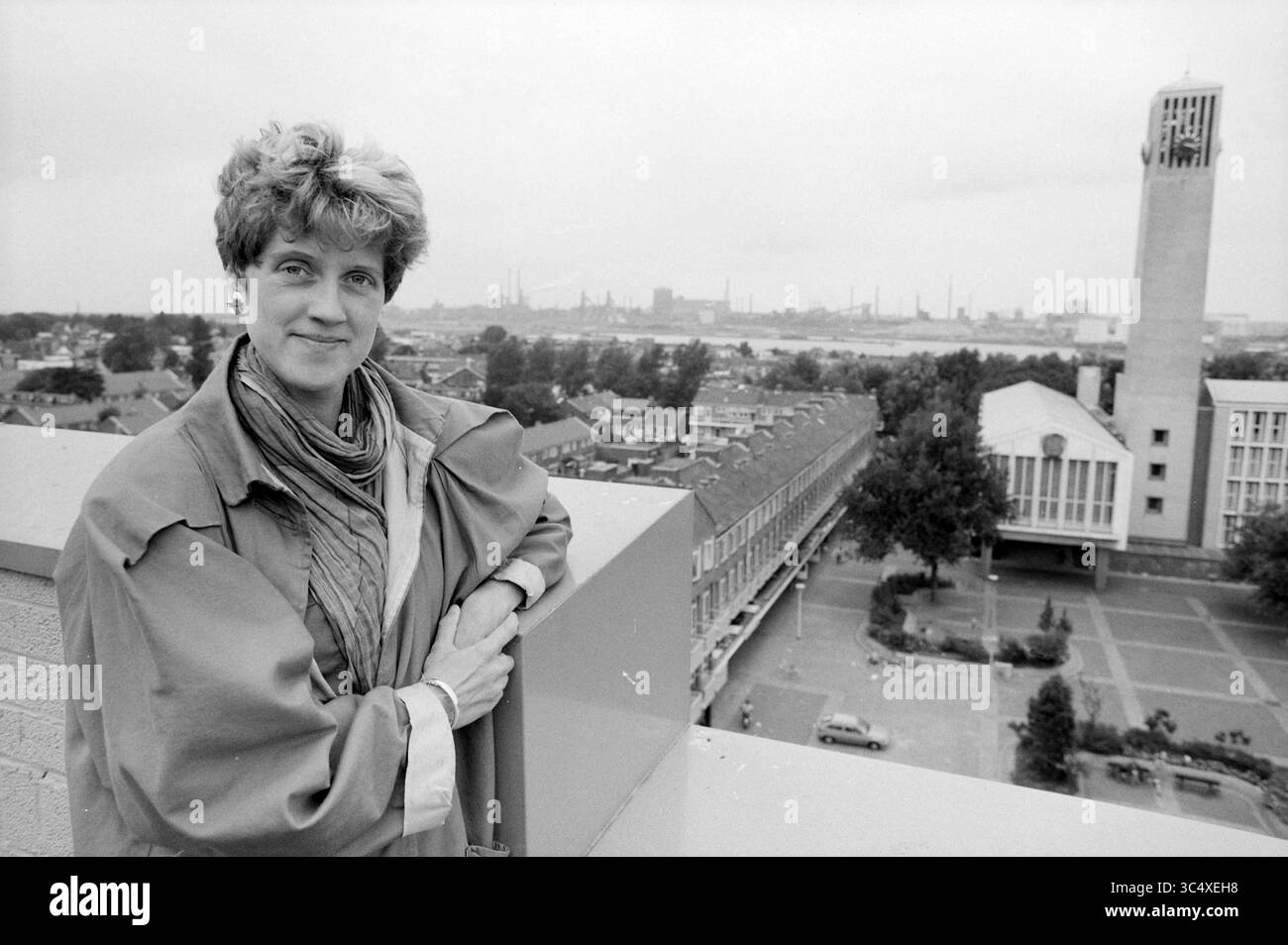 Anne Rietmeijer, Environmental Affairs, IJmuiden, IJmuiden, Niederlande, 22-09-1991 Whizgle News, Dutch Desk, Niederlande, 1950-2000 Eine Frau steht selbstbewusst auf einem Dach und blickt auf eine Stadtlandschaft mit einem prominenten Uhrenturm und einem Raster von Wohngebäuden. Die Szene vermittelt ein Gefühl des urbanen Lebens, wobei ferne Industriestrukturen auf die wirtschaftliche Aktivität der Region hinweisen. Die Kleidung und der Ausdruck der Frau deuten auf eine Mischung aus Professionalität und Zugänglichkeit hin. Stockfoto
