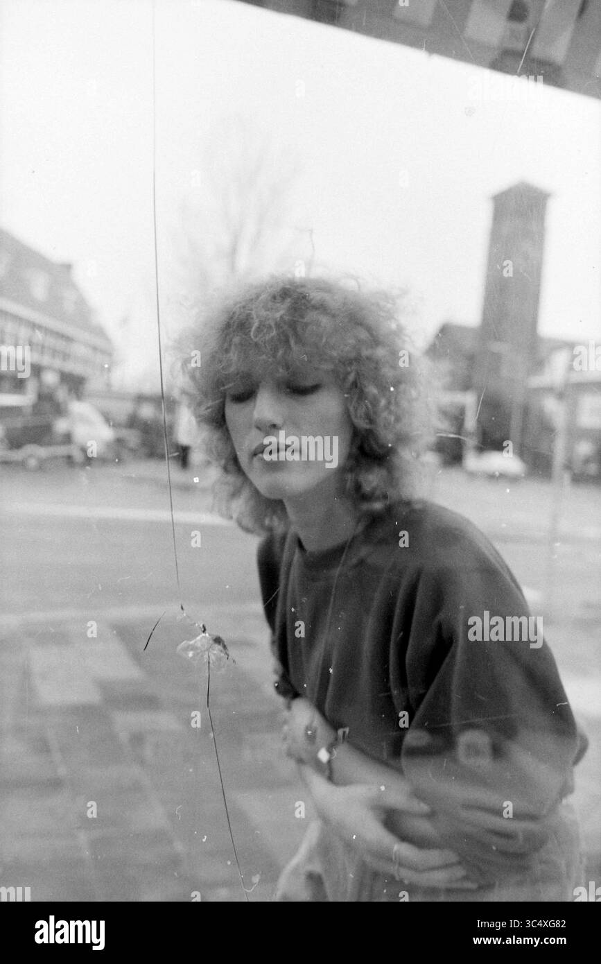 Kugelloch im Schaufenster, Buntglas, Glasblasen, Glas im Allgemeinen, 28-12-1983 Whizgle News, Dutch Desk, Niederlande, 1950-2000 Ein junger Mensch mit lockigen Haaren lehnt sich an eine Glasoberfläche und blickt nachdenklich nach unten. Die Szene draußen zeigt einen Hauch von urbanem Leben mit verschwommenen Umrissen von Gebäuden und Fahrzeugen im Hintergrund. Stockfoto