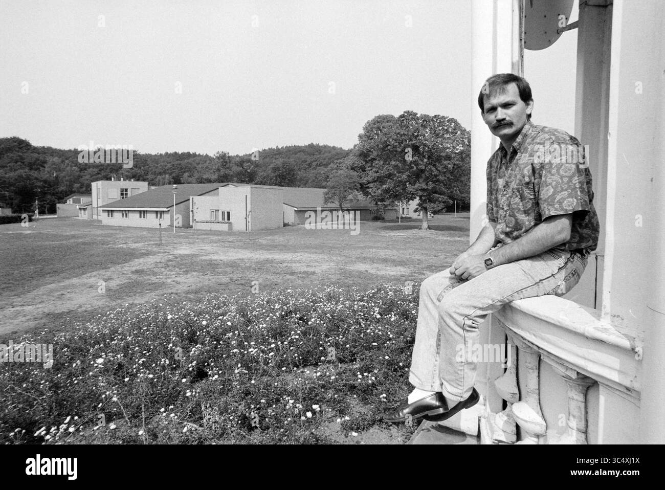 Evertskliniek P.Z., Santpoort, 20-07-1990 Whizgle News, Dutch Desk, Niederlande, 1950-2000 Ein Mann sitzt auf einem Felsvorsprung und blickt in die Ferne, mit einer ruhigen Landschaft aus offenen Feldern und Gebäuden im Hintergrund. Er trägt ein gemustertes Hemd und eine lässige Jeans, die einen Moment der Ruhe in einer ruhigen Umgebung verkörpern. Stockfoto