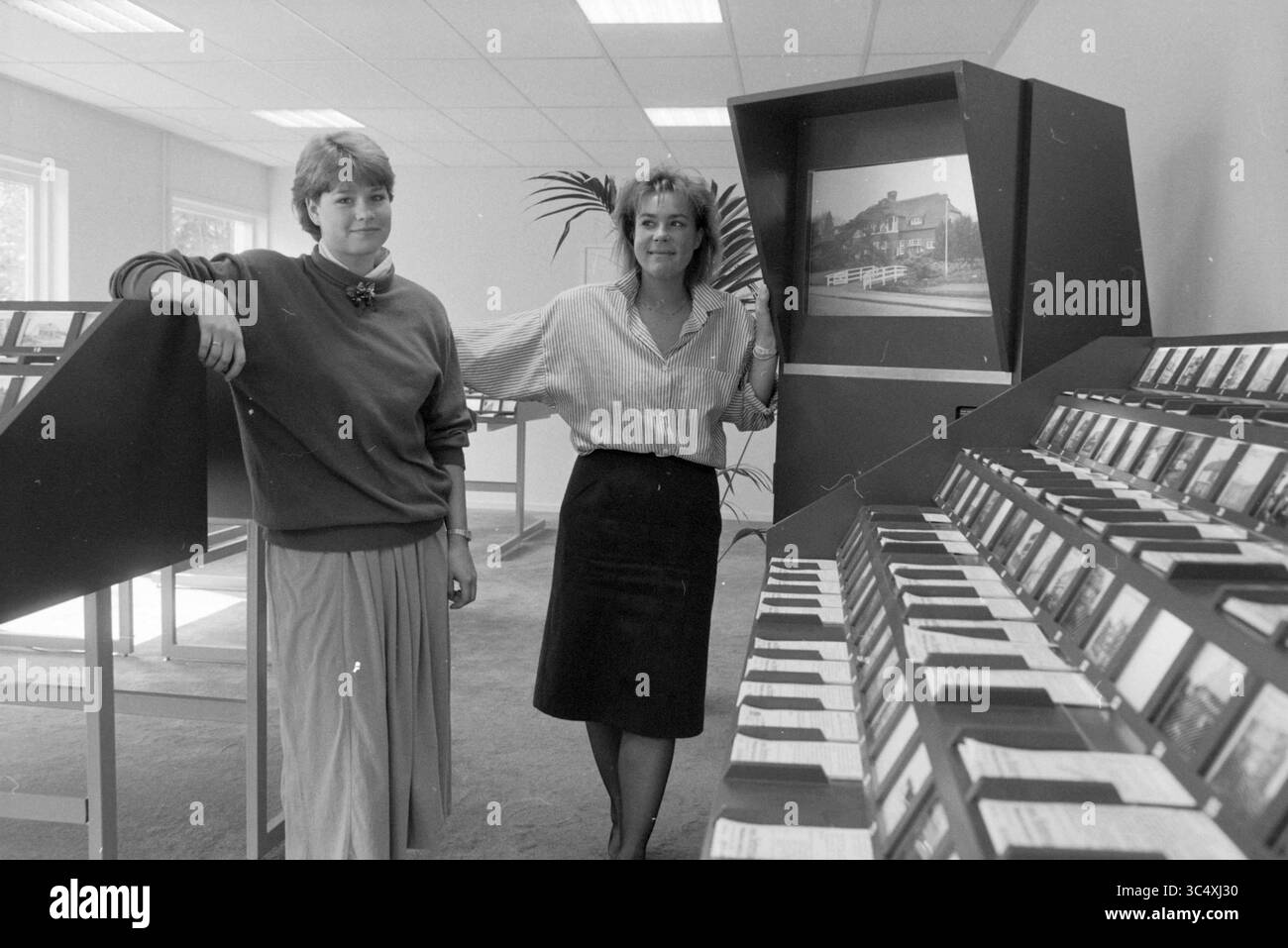 Digitales Angebot von Häusern zum Verkauf, 00-05-1988 Whizgle News, Dutch Desk, Niederlande, 1950-2000 zwei Frauen stehen neben einer Präsentation von ordentlich organisierten Folien, eine lächelt, die andere lehnt sich beiläufig gegen die Anzeige. Sie befinden sich in einem hellen, luftigen Raum, der mit Pflanzen geschmückt ist, was auf eine kreative oder lehrreiche Umgebung hindeutet. Stockfoto