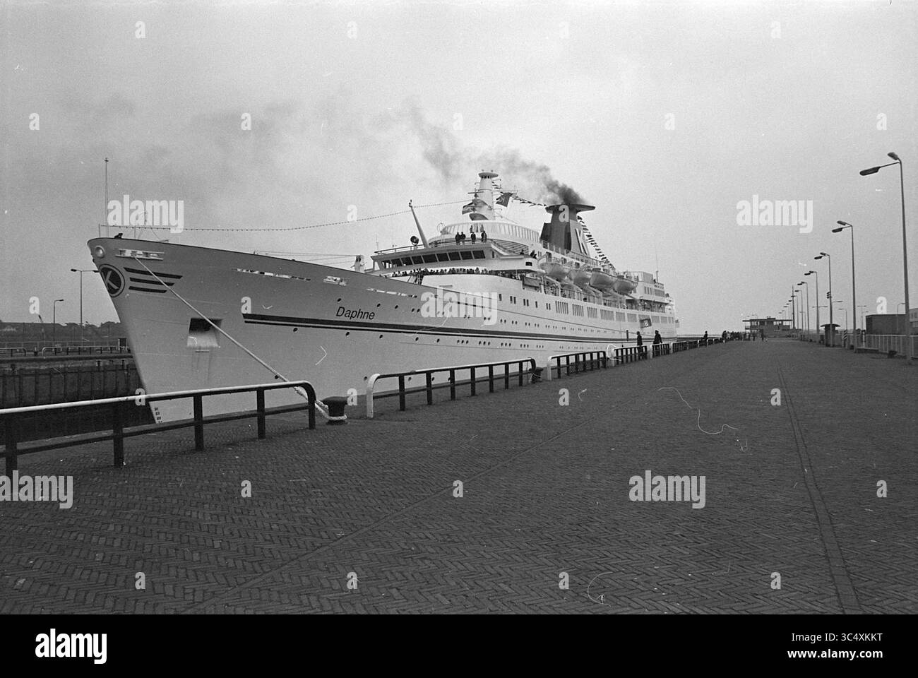 Kreuzfahrtschiff 'The Daphne', Schiffe, 28-05-1976 Whizgle News, Dutch Desk, Niederlande, 1950-2000 Ein majestätisches Kreuzfahrtschiff namens „Daphne“ legt an einem geschäftigen Pier an und raucht aus seinem Trichter. Die Passagiere spazieren entlang der Promenade und genießen die maritime Szene, während die Leuchttürme den Weg zum Schiff säumen. Stockfoto