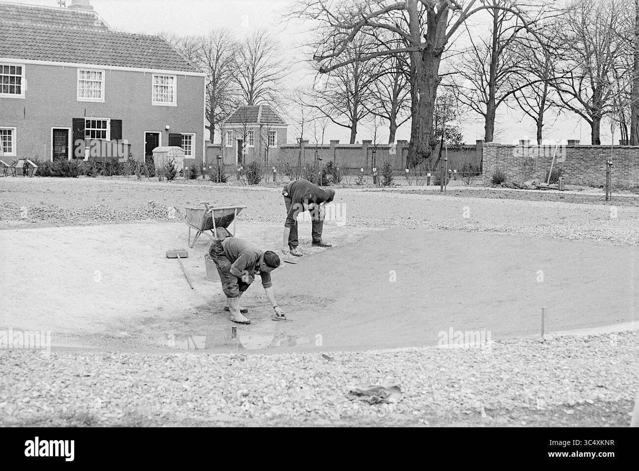 Bau des Beeckestijn-Brunnens. Arbeit, 19-04-1971 Whizgle News, Dutch Desk, Niederlande, 1950-2000 zwei Arbeiter arbeiten im Landschaftsbau, einer kniet, um den Boden zu glätten, während der andere eine Schubkarre in der Nähe benutzt. Um sie herum befinden sich Bäume und ein Wohngebäude im Hintergrund, was auf eine Umgebung hinweist, die sich auf Gartenarbeit und Gartenarbeit konzentriert. Stockfoto