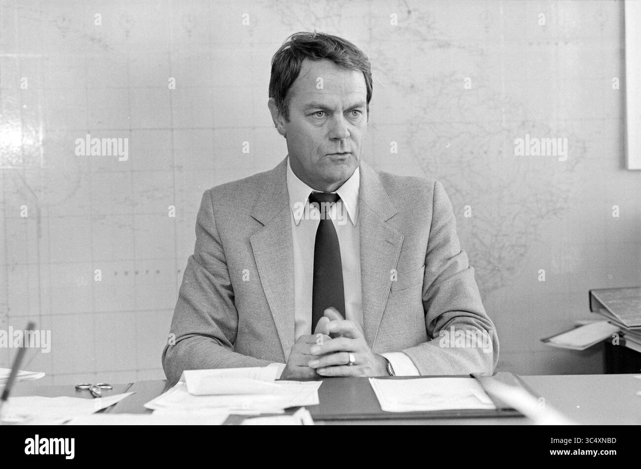Mr. Stuart, Himalaya-Expedition, Expedition, 27-10-1982 Whizgle News, Dutch Desk, Niederlande, 1950-2000 Ein Mann mittleren Alters in einem Anzug sitzt an einem Schreibtisch, seine Hände sind zusammengeschlossen, mit einem fokussierten Ausdruck. Hinter ihm ziert eine Karte die Wand, die auf eine professionelle Umgebung hindeutet. Papier und ein Stift sind über den Schreibtisch verteilt, was auf eine geschäftige Arbeitsumgebung hinweist. Stockfoto