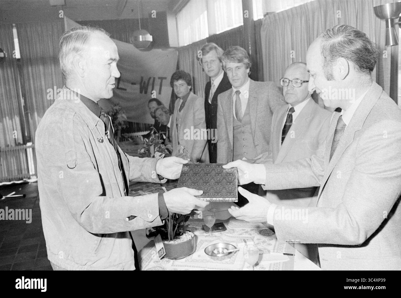 Fußballempfang 60 Jahre Gelb weiß, Empfänge, Fußball, 02-05-1980 Whizgle News, Dutch Desk, Niederlande, 1950-2000 Eine Gruppe von Männern steht an einem Tisch und führt einen bedeutenden Austausch durch. Ein Mann überreicht einem anderen eine dekorative Box, während mehrere andere die Interaktion beobachten und einen wichtigen Moment oder eine Feier in einem formellen Rahmen vorschlagen. Stockfoto