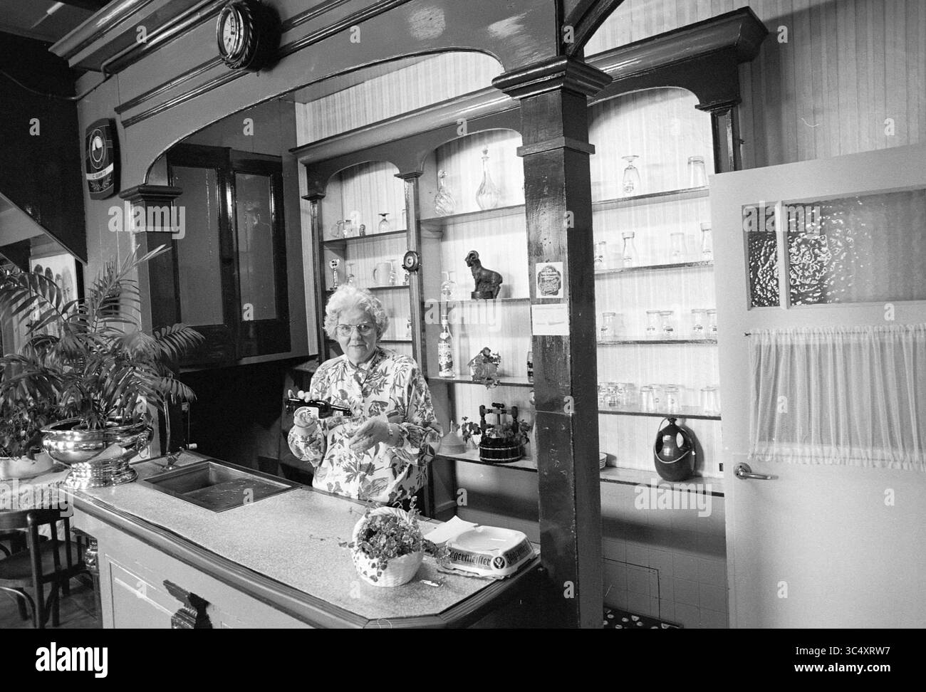 Femke Gerritse, Pub, Driehuis, Driehuis, 24-09-1992 Whizgle News, Dutch Desk, Niederlande, 1950-2000 eine ältere Frau steht hinter einer Bar im Vintage-Stil, umgeben von Regalen mit verschiedenen Glaswaren und Dekorationen. Sie scheint konzentriert zu sein, während sie sich um ihre Arbeit kümmert, und strahlt ein Gefühl von Wärme und Nostalgie in der gemütlichen Umgebung aus. Stockfoto