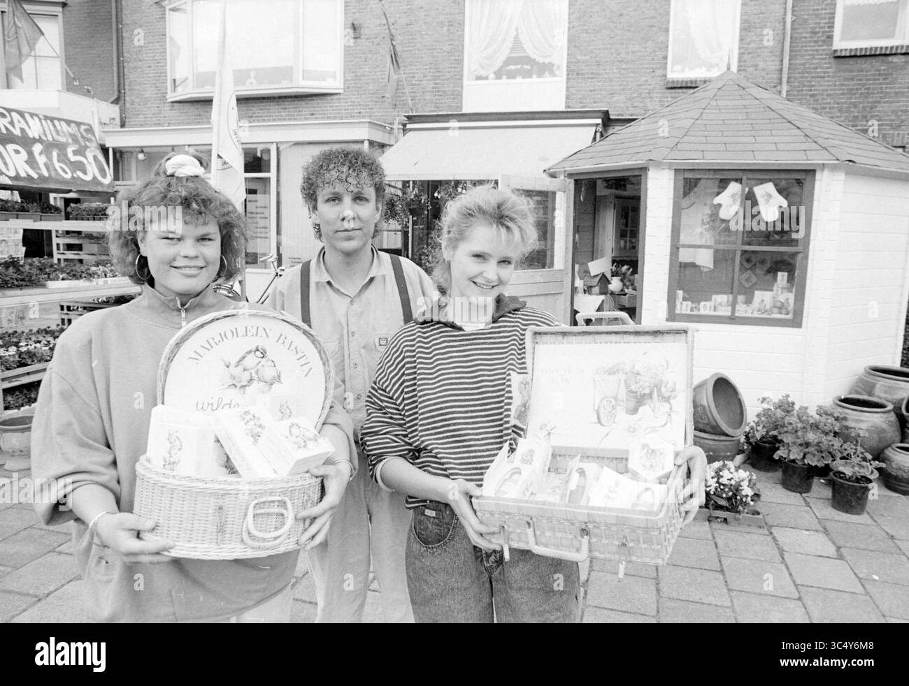 Blumenkunst Monique Henk und Marjon, Blumen und Blumenmädchen, 20-05-1988 Whizgle News, Dutch Desk, Niederlande, 1950-2000 drei Personen stehen draußen, jeder hält Körbe mit verpackten Waren. Sie befinden sich vor einer Schaufensterfront, die mit verschiedenen dekorativen Gegenständen geschmückt ist. Der Hintergrund bietet eine geschäftige Marktatmosphäre, mit Pflanzen und Außenanlagen, die die lebhafte Szene unterstreichen. Stockfoto