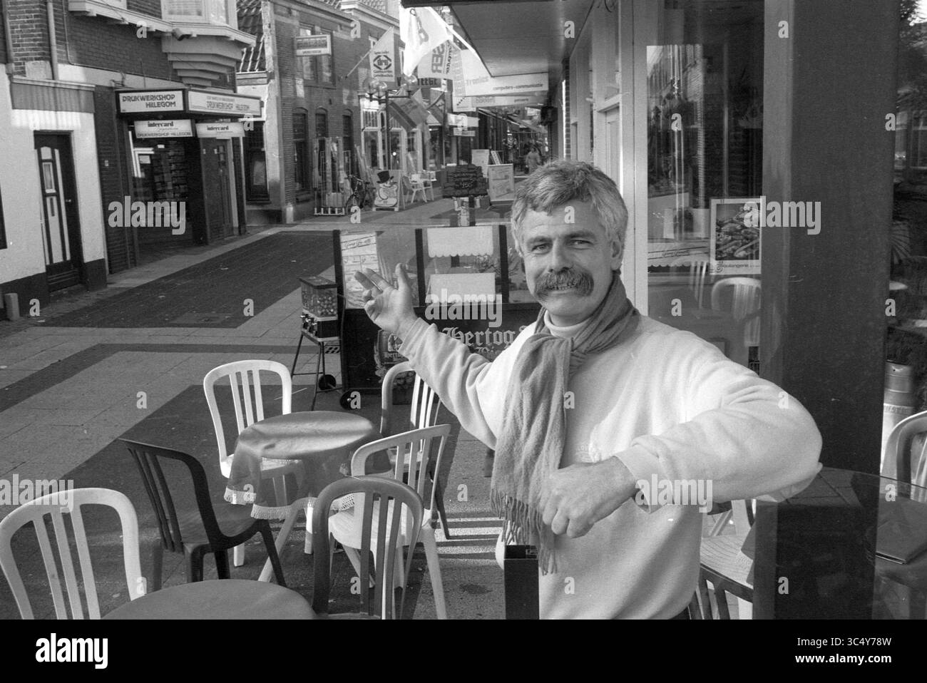 Frans van Houten Coffee Shop Hillegom, Coffee, 30 11.11.1989 Whizgle News, Dutch Desk, Niederlande, 1950-2000 Ein Mann gibt sich vom Bürgersteig vor einem Café aus warm und zeigt die malerische Straße, die von Geschäften und leeren Stühlen gesäumt ist. Er trägt einen leichten Pullover und einen Schal und strahlt eine einladende Atmosphäre in einer charmanten, friedlichen Atmosphäre aus. Stockfoto