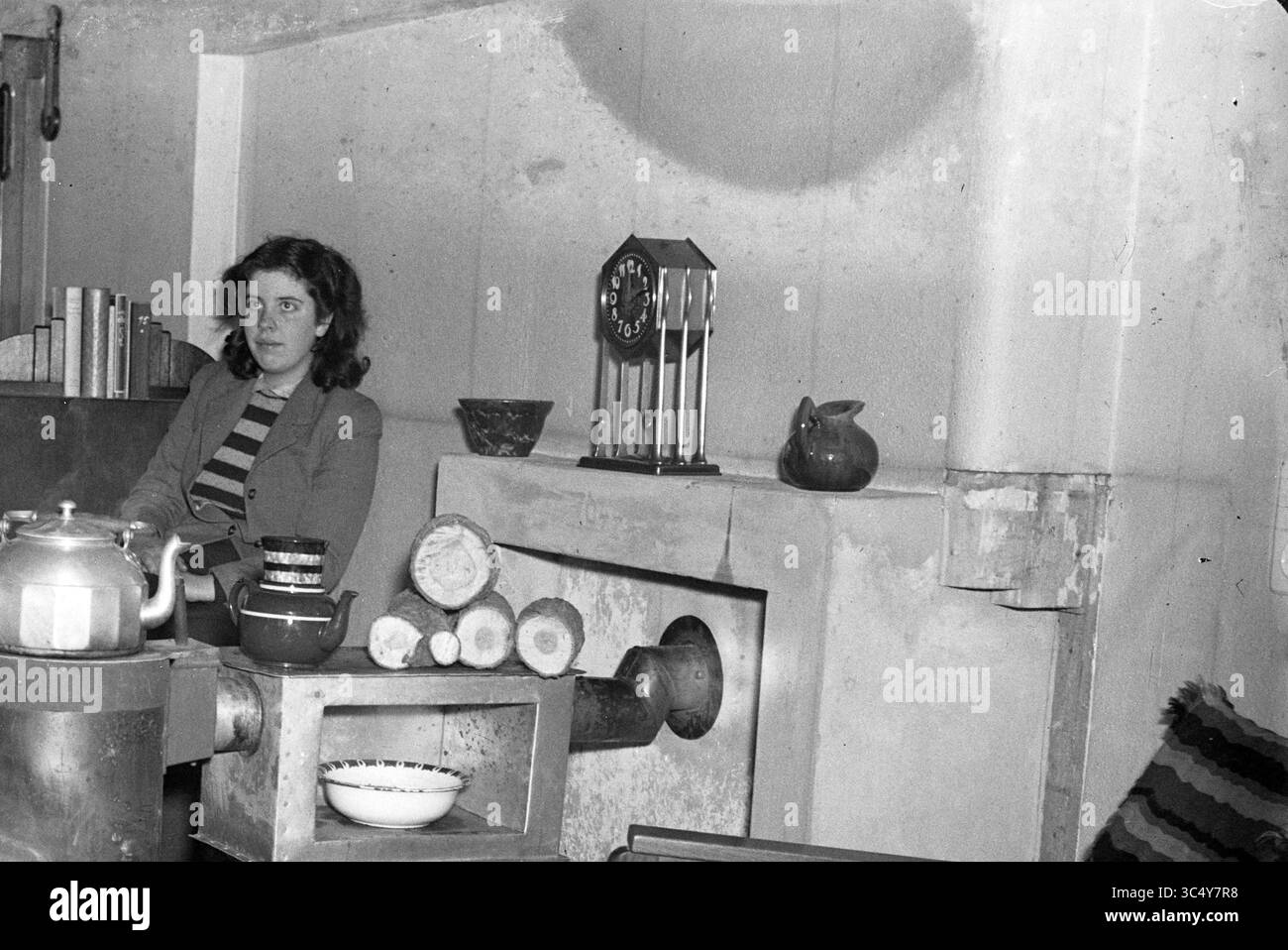 Wohnzimmer mit Kaltbauchofen, 00-00-1951 Whizgle News, Dutch Desk, Niederlande, 1950-2000 Eine junge Frau sitzt nachdenklich in einem gemütlichen Zimmer, umgeben von Büchern und verschiedenen Dekorationsgegenständen, darunter eine Uhr und ein Keramikkaninchen. Ein Herd mit ordentlich gestapelten Baumstämmen trägt zur Wärme des Raumes bei. Stockfoto