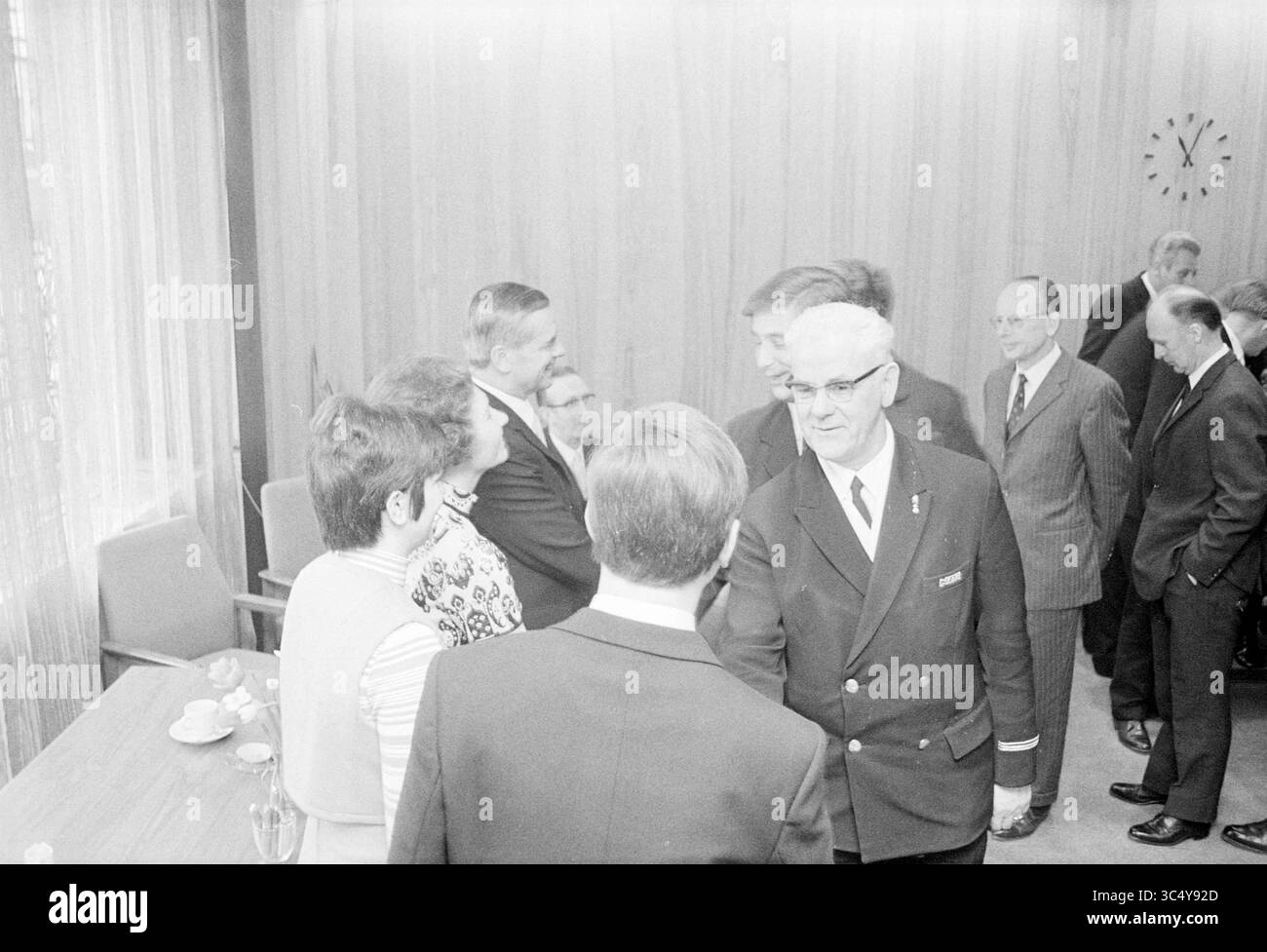 Die Menschen werden bei einem Empfang gratuliert Whizgle News, Dutch Desk, Niederlande, 1950-2000 Eine Gruppe von formell gekleideten Personen führt in einem Konferenzraum Gespräche, mit einer Mischung aus Lächeln und ernsten Ausdrücken. Die Einrichtung ist mit Holztäfelung und einer Uhr an der Wand ausgestattet, was eine professionelle Atmosphäre anregt. Stockfoto