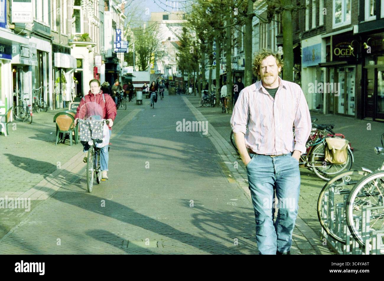 Kees Amama, Haarlem, Niederlande, 01-10-2003 Whizgle News, Dutch Desk, Niederlande, 1950-2000 Eine geschäftige Straßenszene fängt einen Mann ein, der lässig mit Händen in Taschen schlendert, flankiert von Radfahrern und Fußgängern. Geschäfte säumen die Straße und tragen zur lebhaften Atmosphäre bei. Bäume spenden Schatten, während der Tag sich entfaltet. Stockfoto