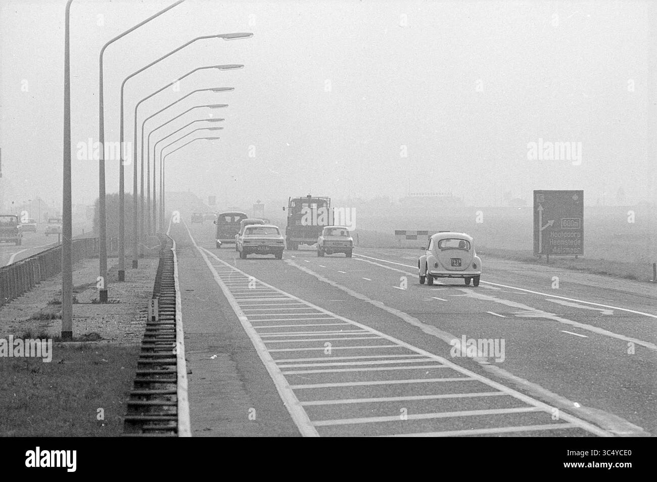Bericht Rijksweg 4 (jetzt A4), Straßen, Straßenbau, Straßenpflanzung, Bericht, 12-10-1970 Whizgle News, Dutch Desk, Niederlande, 1950-2000 Eine ruhige Straße zieht sich in die Ferne, gesäumt von Straßenlaternen. Mehrere Autos fahren entlang der Autobahn, während ein leichter Dunst in der Atmosphäre auftaucht, was ein ruhiges und doch etwas geheimnisvolles Ambiente schafft. Ein Straßenschild weist auf bevorstehende Wegbeschreibungen oder Wahrzeichen hin. Stockfoto
