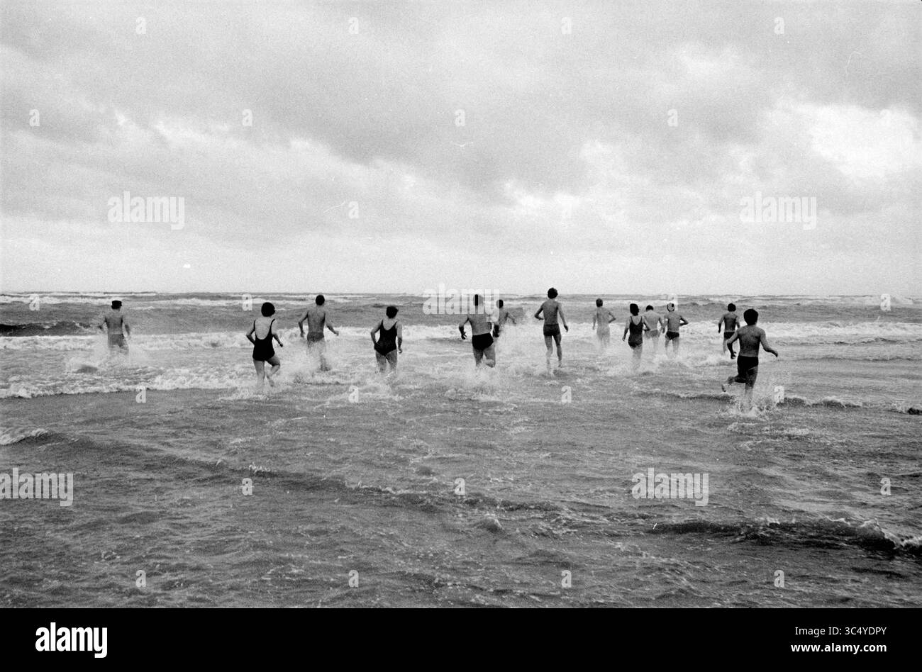 Neujahrstauchgang am Strand von Zandvoort, Zandvoort Whizgle News, Dutch Desk, Niederlande, 1950-2000 Eine Gruppe von Menschen waten in den Ozean und kreieren Spritzer, während sie den Wellen unter einem bewölkten Himmel trotzen. Stockfoto