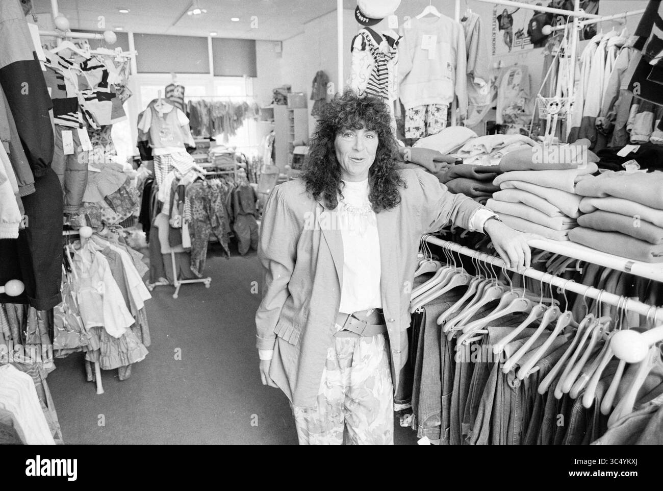 Kindermode Bizbizzy Bloemendaalseweg, Geschäfte und Kioske, Bloemendaal, Bloemendaalseweg, 11-05-1989 Whizgle News, Dutch Desk, Niederlande, 1950-2000 Eine Frau posiert selbstbewusst in einem Bekleidungsgeschäft voller verschiedener Kleidungsstücke, die an Regalen hängen und ordentlich in Regalen gestapelt sind. Das Geschäft bietet eine lebhafte Auswahl an Freizeitkleidung, während sie sich durch ihren einzigartigen Sinn für Mode und lockiges Haar auszeichnet. Stockfoto