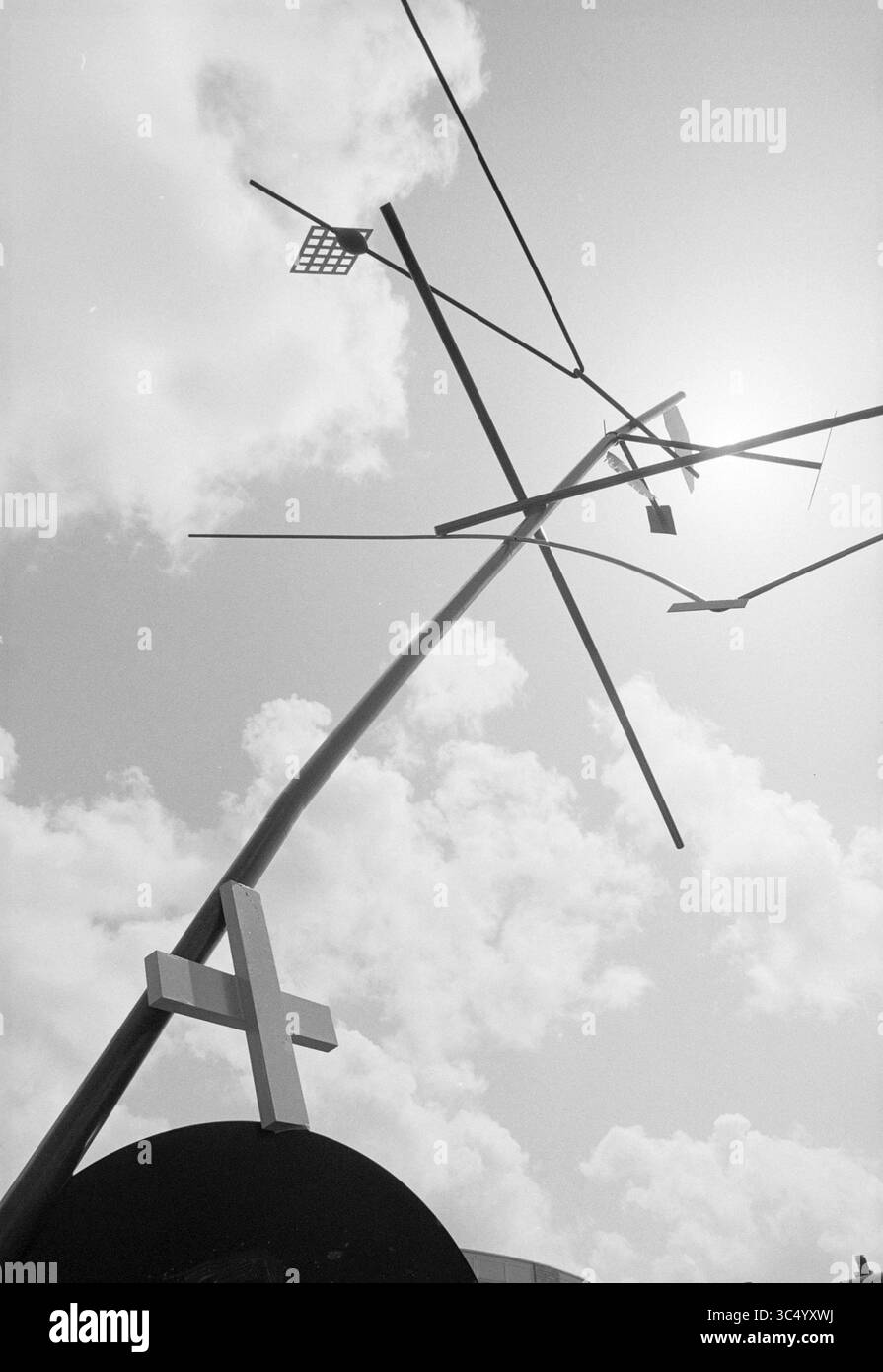 Artwork, Zuiderpolder, 03-05-1994 Whizgle News, Dutch Desk, Niederlande, 1950-2000 Eine hohe, abstrakte Struktur erstreckt sich nach oben, durchkreuzt von Kreuzelementen und reicht bis zu einer hellen Sonne vor dem Hintergrund flauschiger Wolken. Die Komposition weckt ein Gefühl von Spiritualität und moderner Kunst. Stockfoto