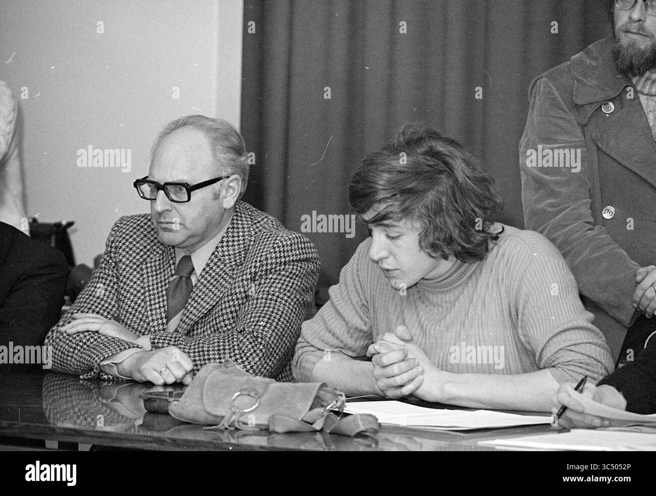 Aktionen bei CIOS Sporttraining bei Duinlust Whizgle News, Dutch Desk, Niederlande, 1950-2000 zwei Personen sitzen während eines Meetings tief in Gedanken an einem Tisch. Der eine trägt eine Brille und einen karierten Anzug und wirkt kontemplativ, während der andere, in einem Pullover gekleidet, die Hände vor sich gefesselt hat und sich auf die Diskussion konzentriert. Eine Gruppe von Personen kann im Hintergrund gesehen werden, was auf eine ernste Atmosphäre hindeutet. Stockfoto