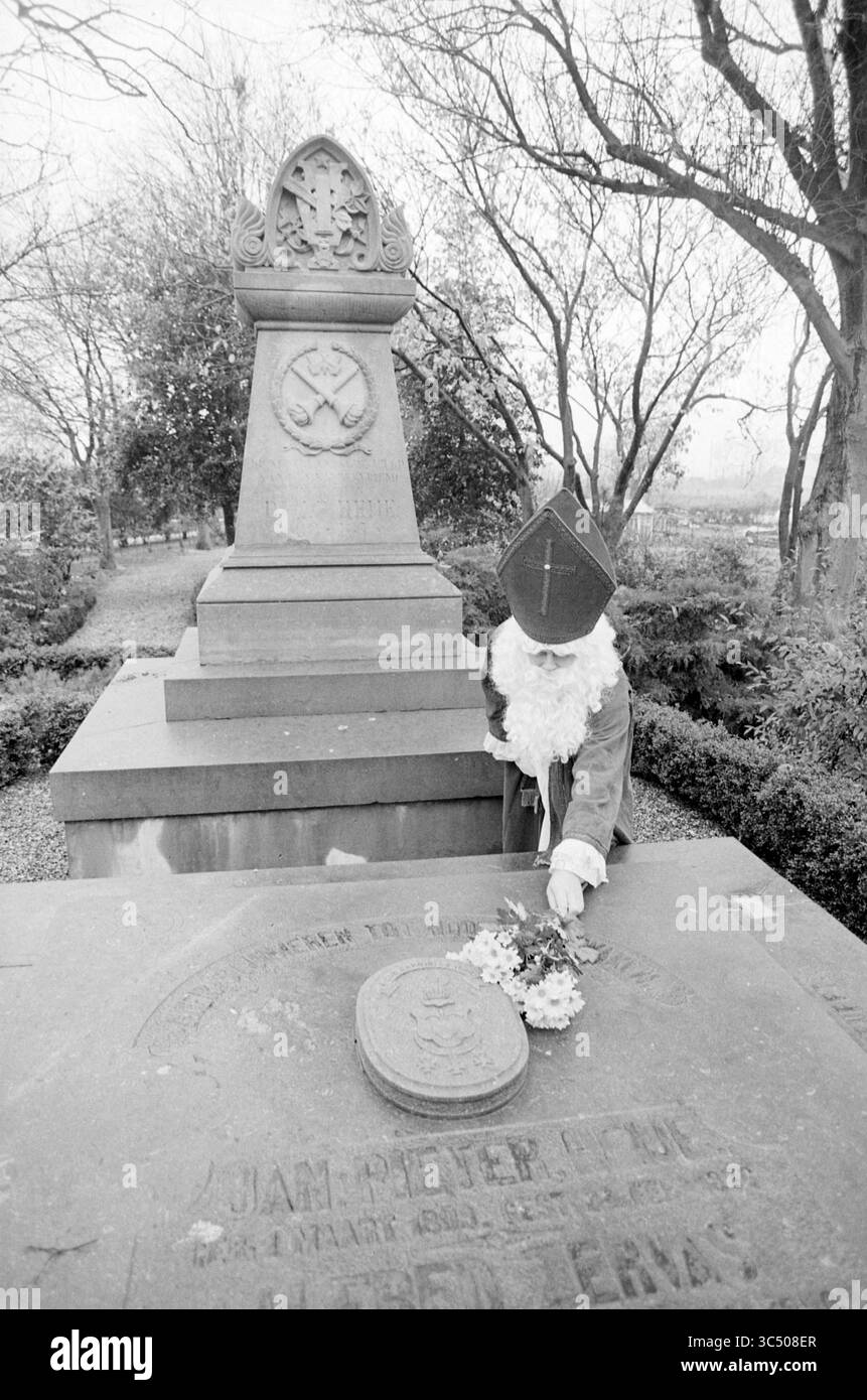Sinterklaas am Grab in Abbenes von J. P. Hey, St. Nikolaus, Sinterklaas, Abbenes, 22-11-1987 Whizgle News, Dutch Desk, Niederlande, 1950-2000 neben einem verwitterten Grabstein steht Eine Figur, die als historische Figur gekleidet ist und sanft Blumen auf den Grabstein legt, inmitten einer ruhigen, von Bäumen gesäumten Umgebung. Die Atmosphäre vermittelt ein Gefühl der Erinnerung und Ehrfurcht. Stockfoto