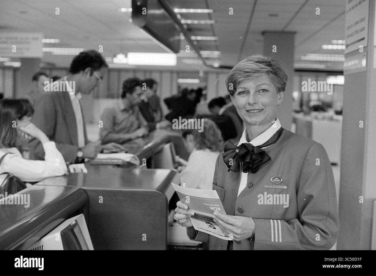 Janny Brevoord, KLM, Schiphol, Schiphol, 14-05-1992 Whizgle News, Dutch Desk, Niederlande, 1950-2000 Eine Flugbegleiterin in intelligenter Uniform lächelt, während sie ein Reisebroschüre in der Hand hält, während Passagiere an einem Check-in-Schalter im Hintergrund anstehen. Stockfoto