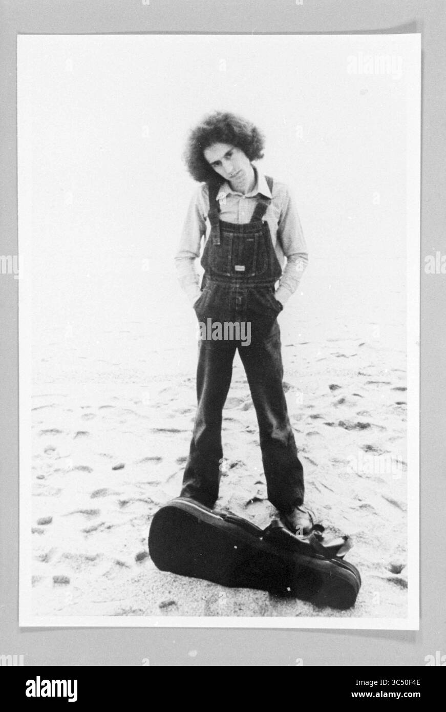 Geiger am Strand, 00-01-1982 Whizgle News, Dutch Desk, Niederlande, 1950-2000 Ein junger Mensch steht auf einer sandigen Oberfläche, trägt Overall und ein Button-up-Hemd. Sie haben lockiges Haar und einen nachdenklichen Ausdruck, mit einem Gitarrenkoffer neben ihren Füßen. Die Szene vermittelt ein Gefühl von Introspektion und künstlerischer Präsenz. Stockfoto
