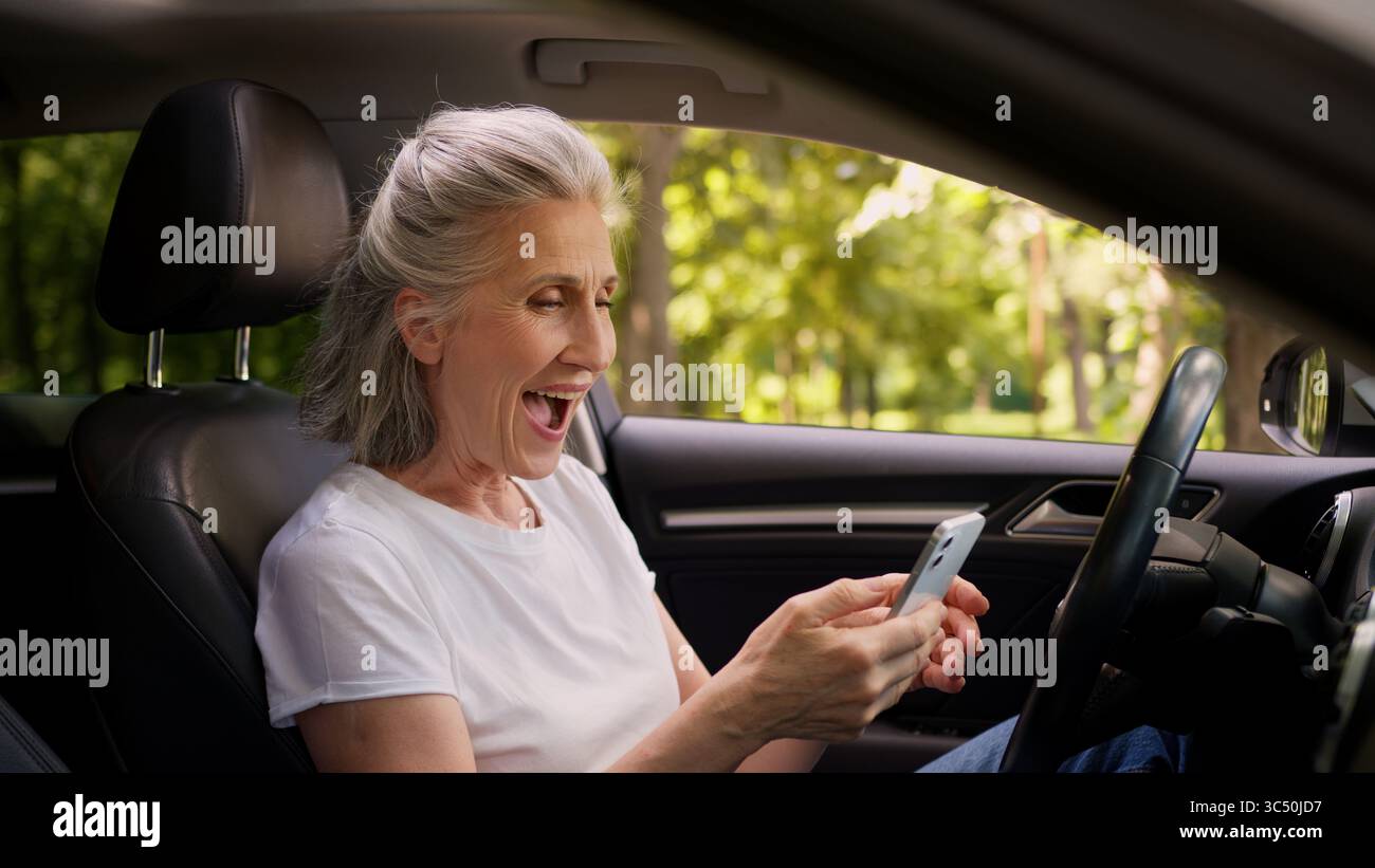 Fröhliche kaukasische reife Frau ältere ältere ältere Frau Rentner Auto Fahrer im Auto mit Handy spielen Spiel Ballen Faust Stockfoto
