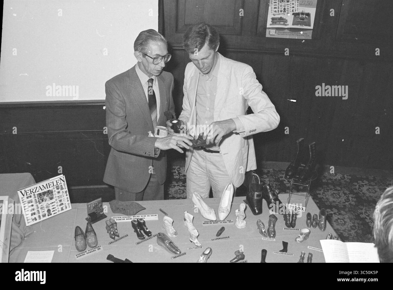 Collector's fair, 02.06.1982 Whizgle News, Dutch Desk, Niederlande, 1950-2000 zwei Männer diskutieren, während sie eine Sammlung verschiedener Schuhmodelle und Artefakte auf einem Tisch betrachten. Ein Mann zeigt ein bestimmtes Stück, während der andere aufmerksam beobachtet. Hinter ihnen ist ein Board mit Text und Bildern sichtbar, das der Sammlung Kontext verleiht. Stockfoto