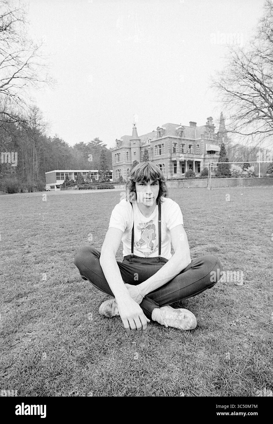 Porträt von Peter Braunsweijger, Portraits, 03-04-1974 Whizgle News, Dutch Desk, Niederlande, 1950-2000 Ein junger Mensch sitzt im Kreuz auf einem grasbewachsenen Rasen, trägt ein grafisches T-Shirt und Hosenträger. Im Hintergrund steht ein beeindruckendes Herrenhaus, umgeben von Bäumen, was eine ruhige und dennoch großartige Umgebung suggeriert. Stockfoto