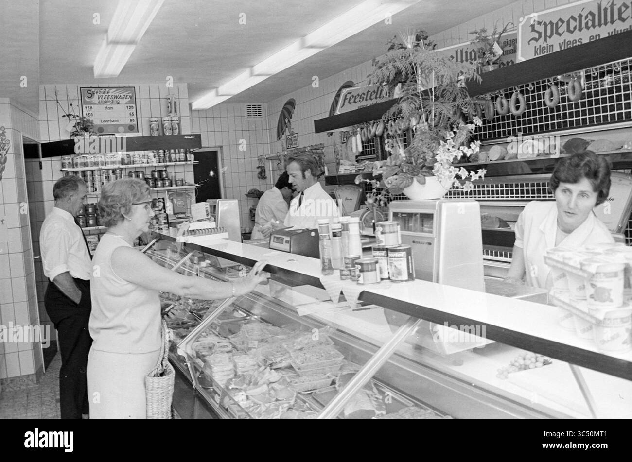 Innenraum einer Metzgerei Whizgle News, Dutch Desk, Niederlande, 1950-2000 In Einem belebten Feinkostgeschäft befindet sich eine Frau an der Theke, die sich mit einem Mitarbeiter unterhalten, während zwei Männer im Hintergrund durch die Regale schauen. Die Ausstellungen zeigen eine Vielzahl von Fleisch- und verpackten Waren, umgeben von lebendigen Pflanzen und fröhlichen Schildern. Stockfoto