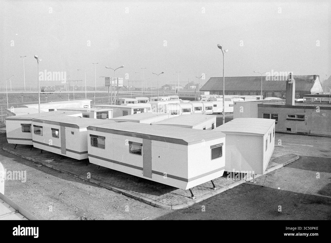 Standort mit Mobilheimen, 00-00-1972 Whizgle News, Dutch Desk, Niederlande, 1950-2000 Eine Sammlung geparkter Mobilheime, die in Reihen angeordnet sind, mit umgebenden Straßenlaternen und Industriebauten im Hintergrund, die einen temporären Wohnraum oder Campingplatz vorschlagen. Stockfoto
