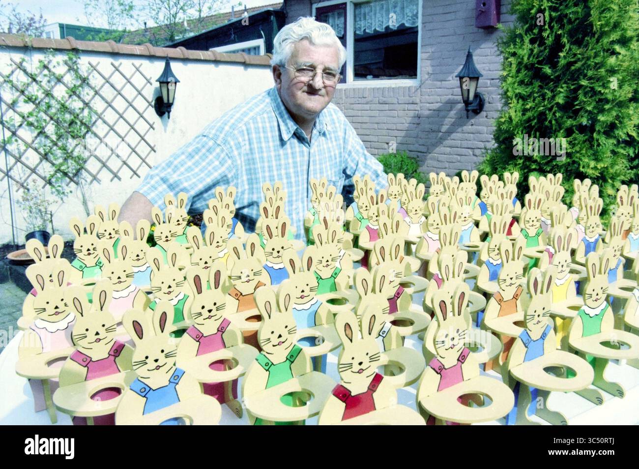 Mr. Van Schooten mit Osterhasen, 16-04-2000 Whizgle News, Dutch Desk, Niederlande, 1950-2000 ein älterer Mann steht stolz inmitten einer Vielzahl von bunten Stühlen mit Häschen-Motiven, von denen jeder verspielte Karikaturkaninchen trägt. Die fröhliche Atmosphäre verleiht der Szene einen skurrilen Touch und unterstreicht ein Gefühl von Nostalgie und Spaß. Stockfoto