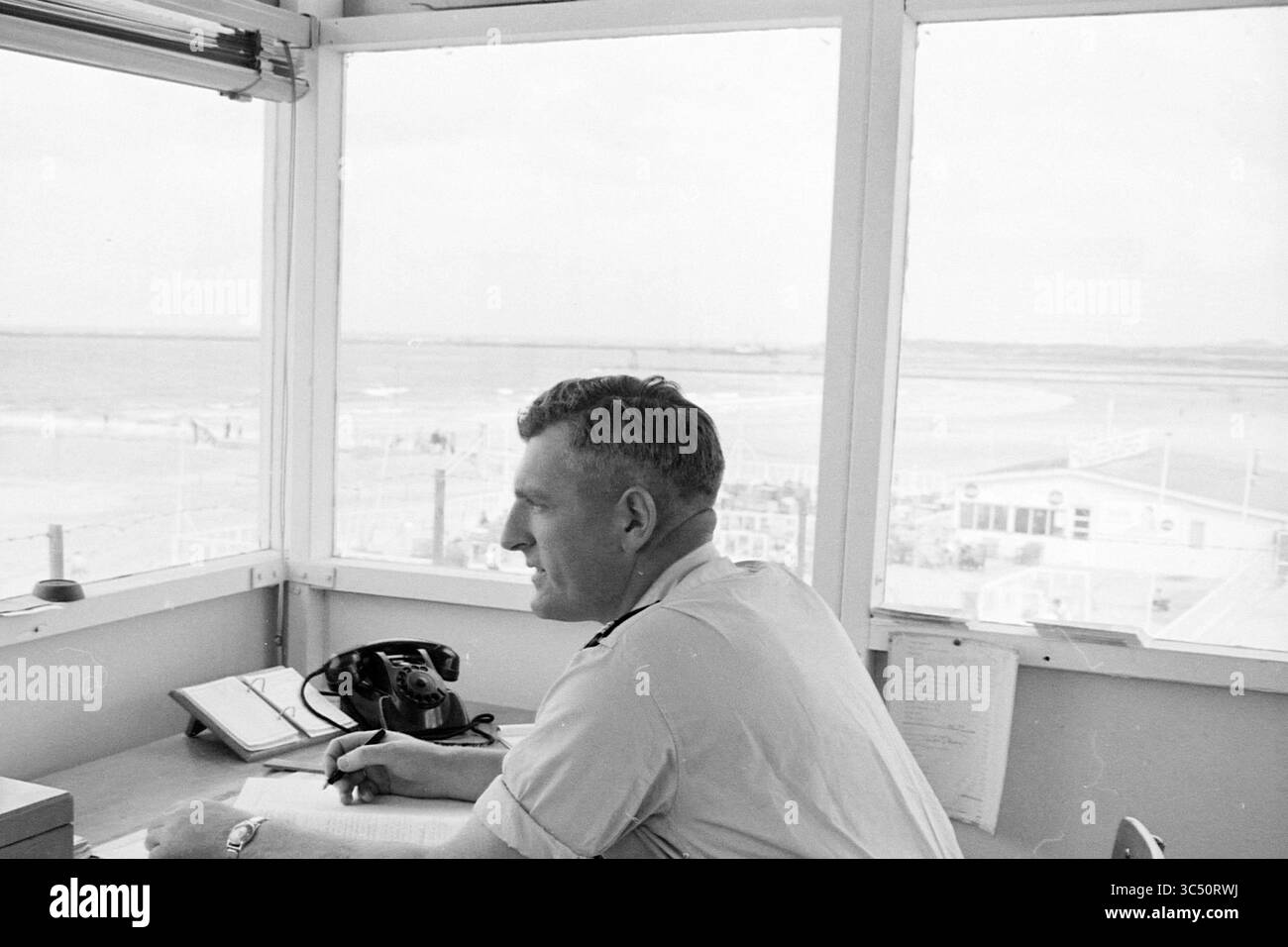 Beach Police IJmuiden, Police, 17-08-1961 Whizgle News, Dutch Desk, Niederlande, 1950-2000 Ein Mann sitzt an einem Schreibtisch in einem Aussichtsturm und blickt auf einen weiten Blick auf die Küste. Er ist mit Schreiben beschäftigt, mit einem altmodischen Telefon auf dem Schreibtisch neben ihm. Die Szene deutet auf eine ruhige, aber aufmerksame Atmosphäre hin, die seine Rolle bei der Überwachung des Strandbereichs im Freien unterstreicht. Stockfoto
