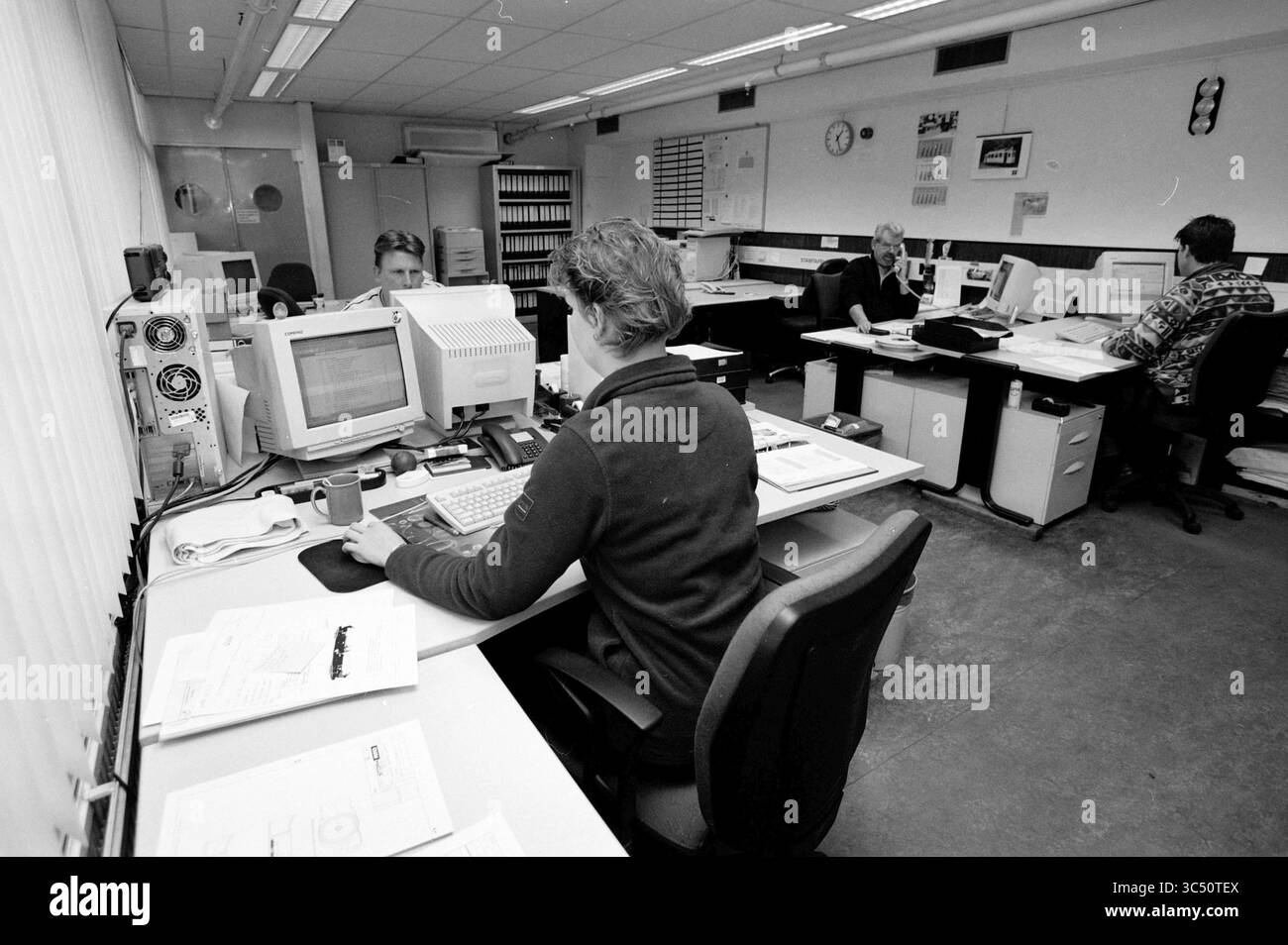 Innenraum eines Büros Whizgle News, Dutch Desk, Niederlande, 1950-2000 Eine geschäftige Büroumgebung, in der mehrere Personen an ihren Schreibtischen arbeiten, die sich auf Computer und Papiere konzentrieren, umgeben von Büroausstattung und Dokumenten. Stockfoto