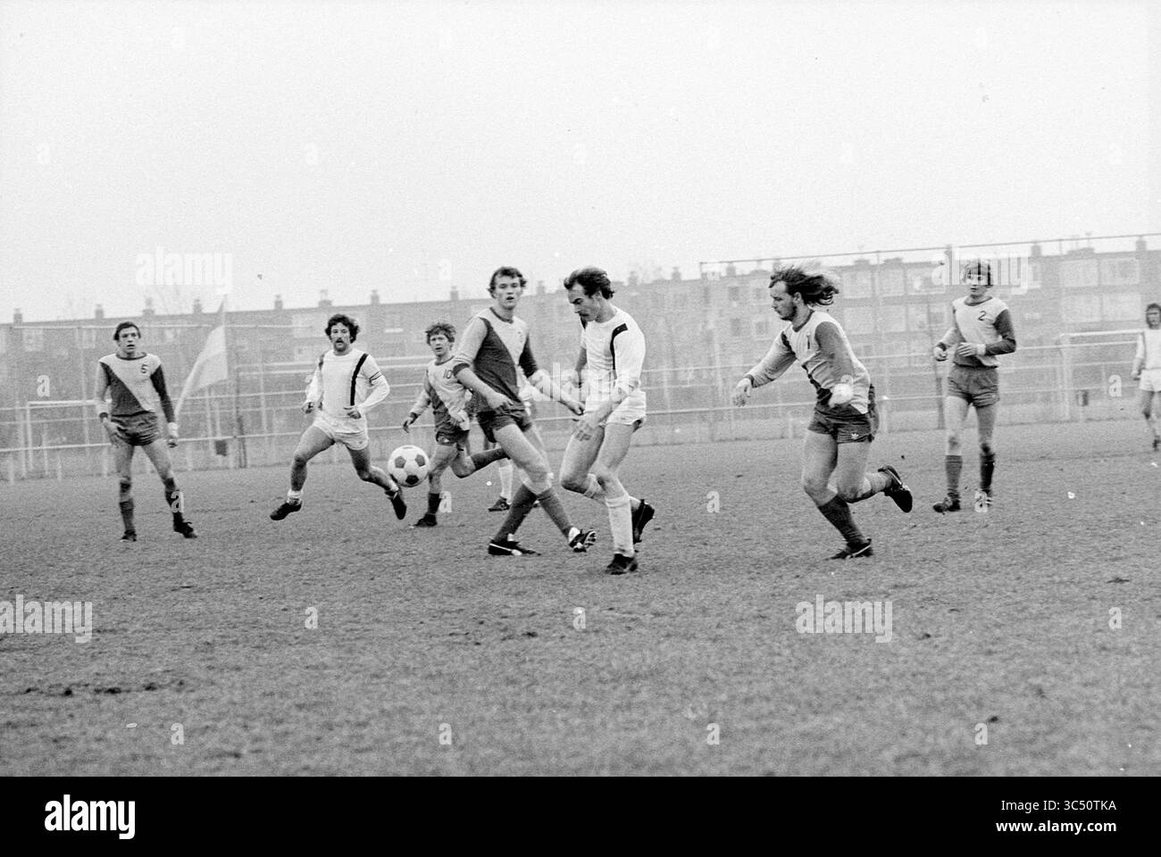 Football Match Whizgle News, Dutch Desk, Niederlande, 1950-2000 Eine Gruppe von Fußballspielern tritt aktiv auf einem grasbewachsenen Feld an und zeigt einen dynamischen Moment im Spiel, während ein Spieler den Ball dribbelt, während andere jagen. Die Kulisse bietet eine Reihe von Gebäuden, die auf eine lebhafte Umgebung hinweisen. Stockfoto