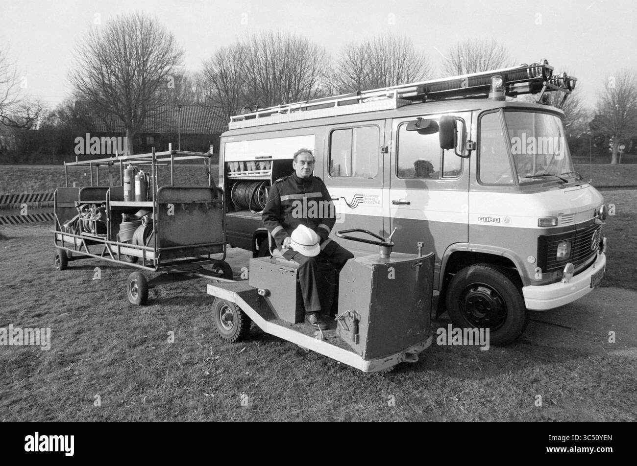Herr van de Heyde, stellvertretender Befehlshaber der Feuerwehr PZ Vogelenzang, Feuer, Feuerwehr, Vogelenzang, 27-01-1989 Whizgle News, Dutch Desk, Niederlande, 1950-2000 Ein Mann sitzt auf einem kleinen, motorisierten Fahrzeug neben einem mit Ausrüstung beladenen Anhänger neben einem alten Feuerwehrwagen. Bäume sind im Hintergrund sichtbar und schaffen eine ruhige Umgebung im Freien. Die Szene fängt einen Moment des Gemeindedienstes oder des Brandschutzes ein. Stockfoto