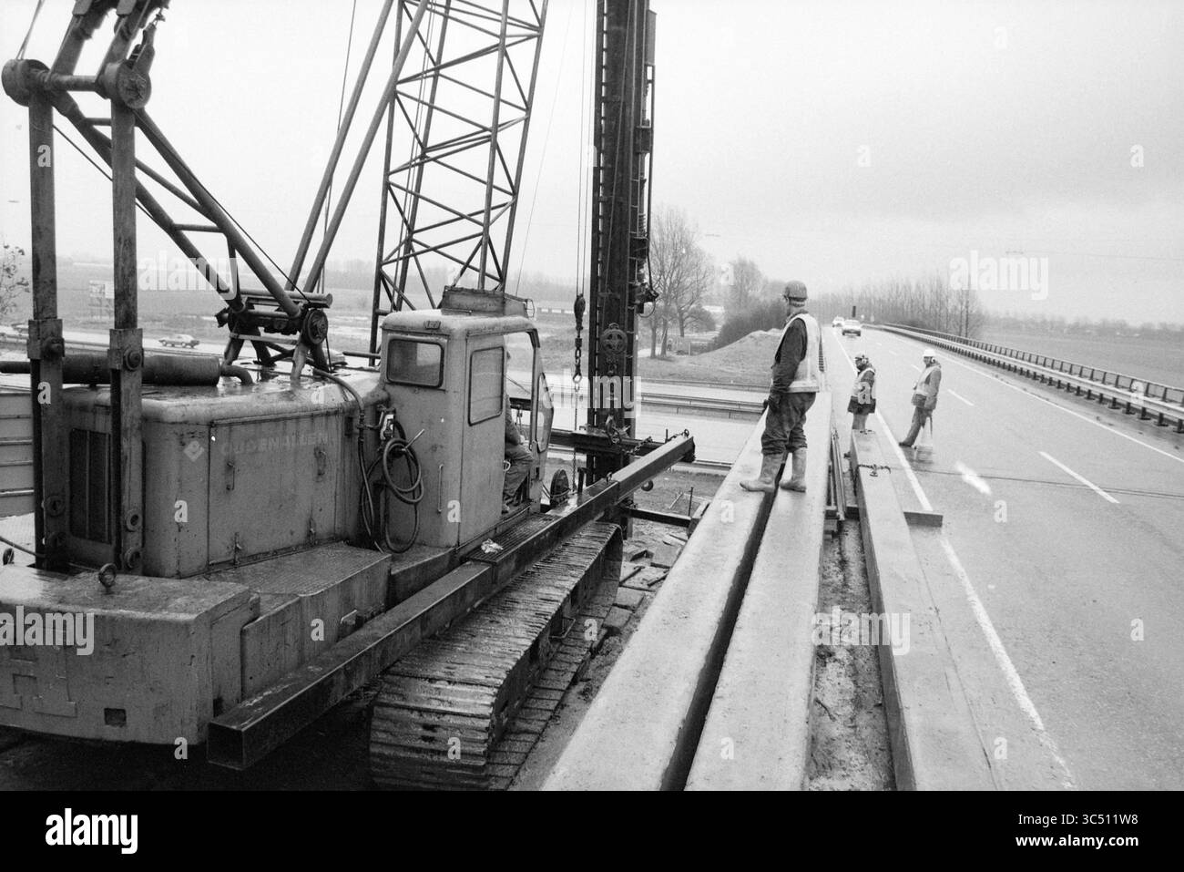 Bau des Radweges Bennebroekerweg, A4, 06-11-1991 Whizgle News, Dutch Desk, Niederlande, 1950-2000 Eine Gruppe von Bauarbeitern beaufsichtigt eine schwere Maschine, die sich neben einer Autobahn befindet, auf der Fertigbetonbalken für eine Brücke aufgestellt werden. In der Ferne sind Fahrzeuge sichtbar, die entlang der Straße fahren, während Bäume den Hintergrund unter einem bewölkten Himmel säumen. Stockfoto