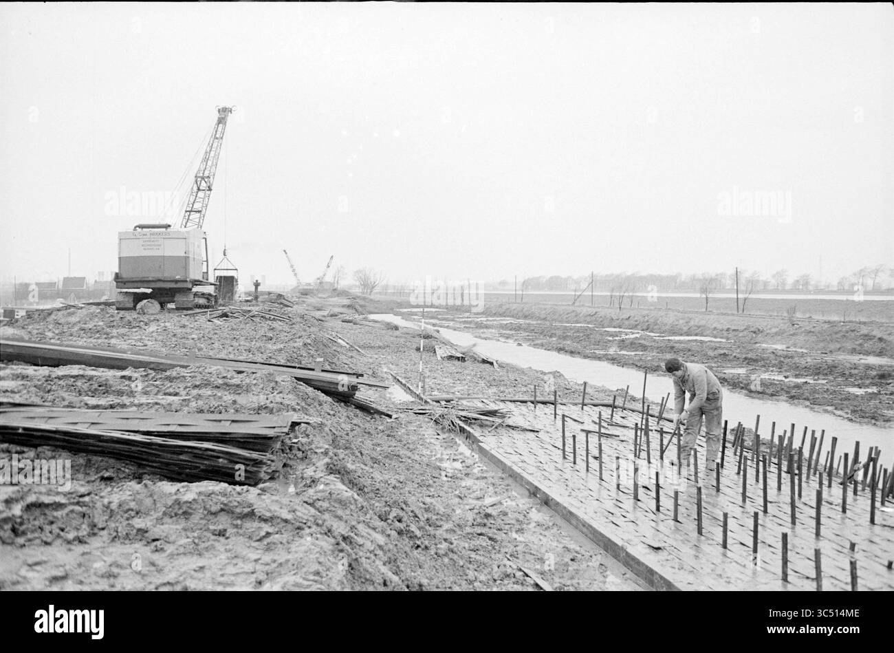 Mann, der Bodenarbeiten mit einem Kran auf der linken Seite macht Whizgle News, Dutch Desk, Niederlande, 1950-2000 Ein Bauarbeiter arrangiert sorgfältig Bewehrungsstäbe auf einer schlammigen Fläche, wobei Maschinen und Kräne im Hintergrund sichtbar sind, was auf die laufende Entwicklung in einem ländlichen Gebiet hinweist. Stockfoto