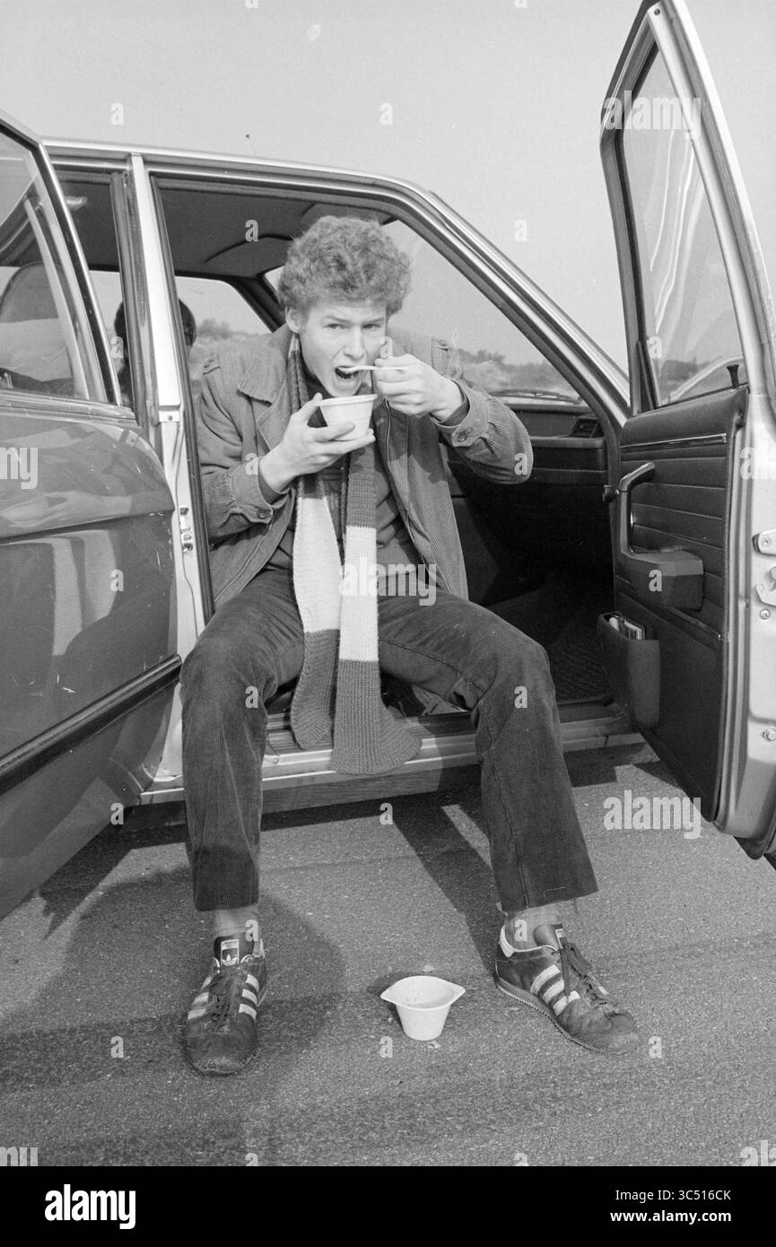 Beach Walk Haarlems Dagblad, Haarlems Dagblad etc. Und IJmuider Courant and Koeri, Walking, 31-07-1980','15-02-1981 Whizgle News, Dutch Desk, Niederlande, 1950-2000 Ein junger Mann sitzt in der offenen Tür eines Autos und genießt eine Mahlzeit aus einer Schüssel, während er einen gestreiften Schal trägt. Er wirkt entspannt und lässig, mit einem verspielten Ausdruck, während er seinen ersten Biss nimmt. Ein Becher sitzt neben ihm auf dem Boden. Stockfoto