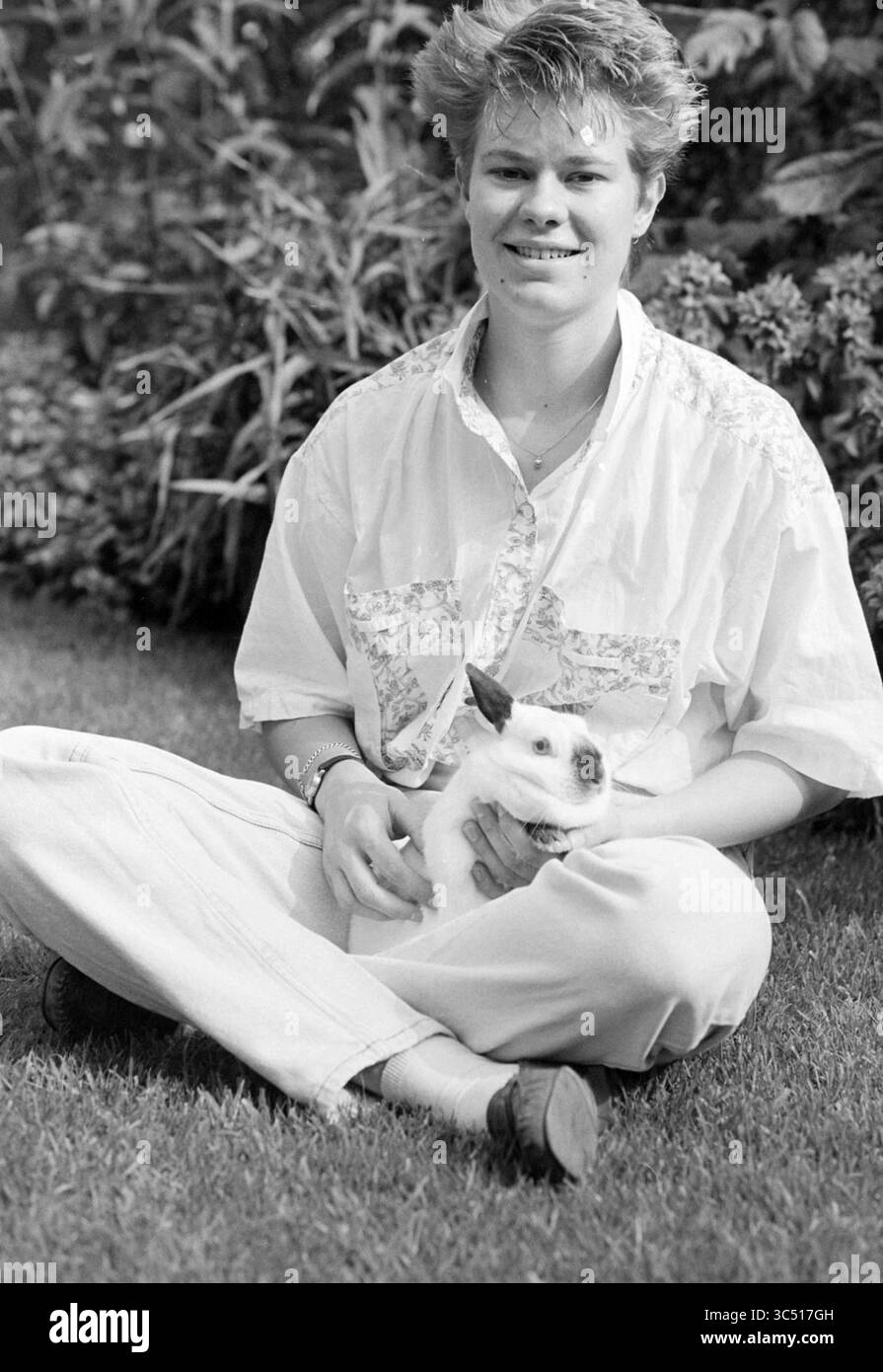 Pass, Karin Jintes, 01-07-1991 Whizgle News, Dutch Desk, Niederlande, 1950-2000 Ein junger Mensch sitzt mit einem fröhlichen Lächeln auf dem Gras und hält sanft ein kleines Kaninchen in den Armen. Das lässige Outfit und die entspannte Haltung ergänzen die ruhige, grüne Umgebung. Stockfoto