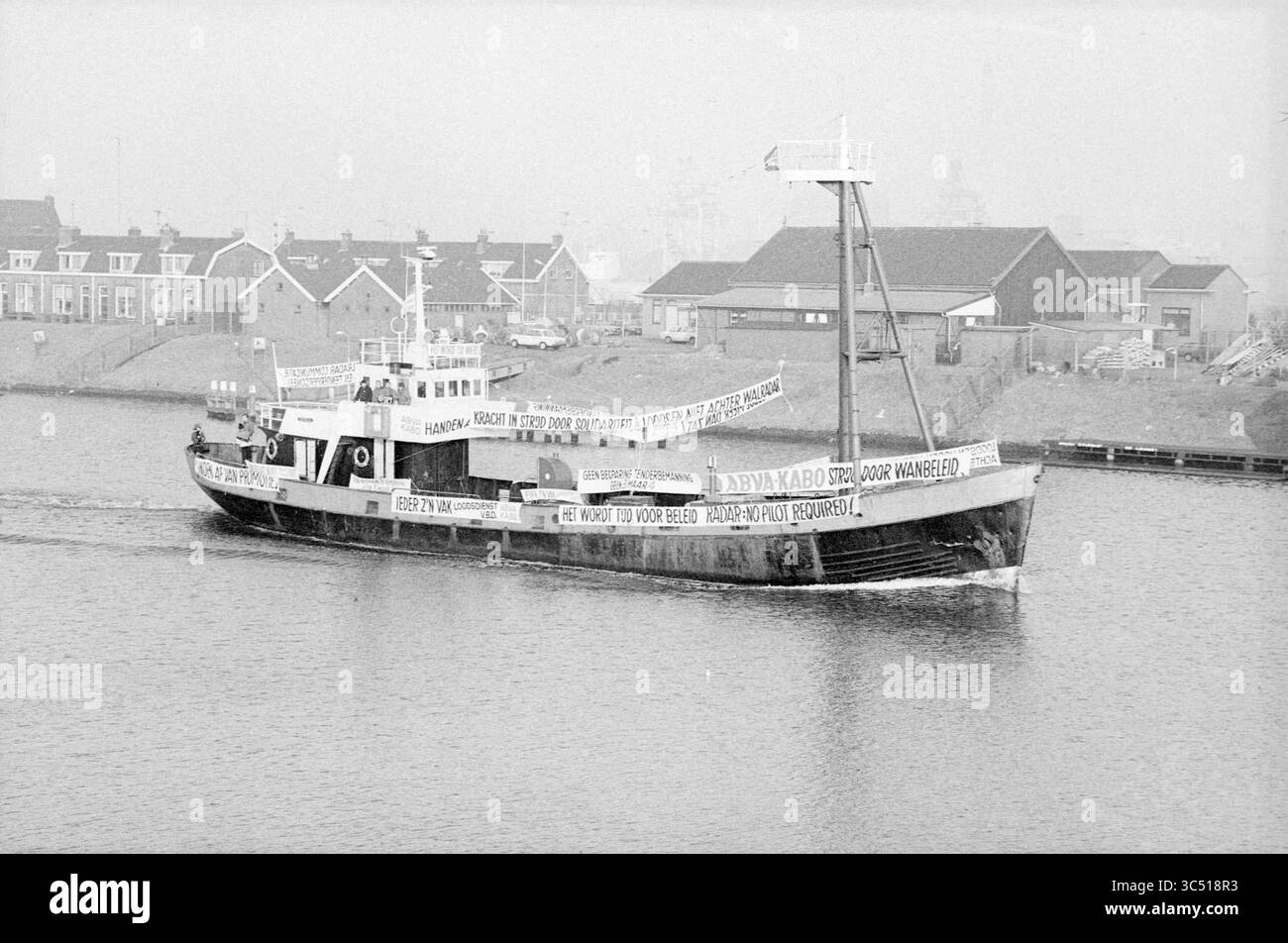 Aktion des Lotsenpersonals, Lotsendienst, Piloten, IJmuiden, Niederlande, 04-02-1982 Whizgle News, Dutch Desk, Niederlande, 1950-2000 Ein Fischerboot navigiert durch ruhige Gewässer, verziert mit großen Bannern mit Botschaften und Slogans. In der Nähe liegen malerische Häuser entlang der Küste, die ein ruhiges Ambiente schaffen, während das Boot vorbeifährt. Stockfoto
