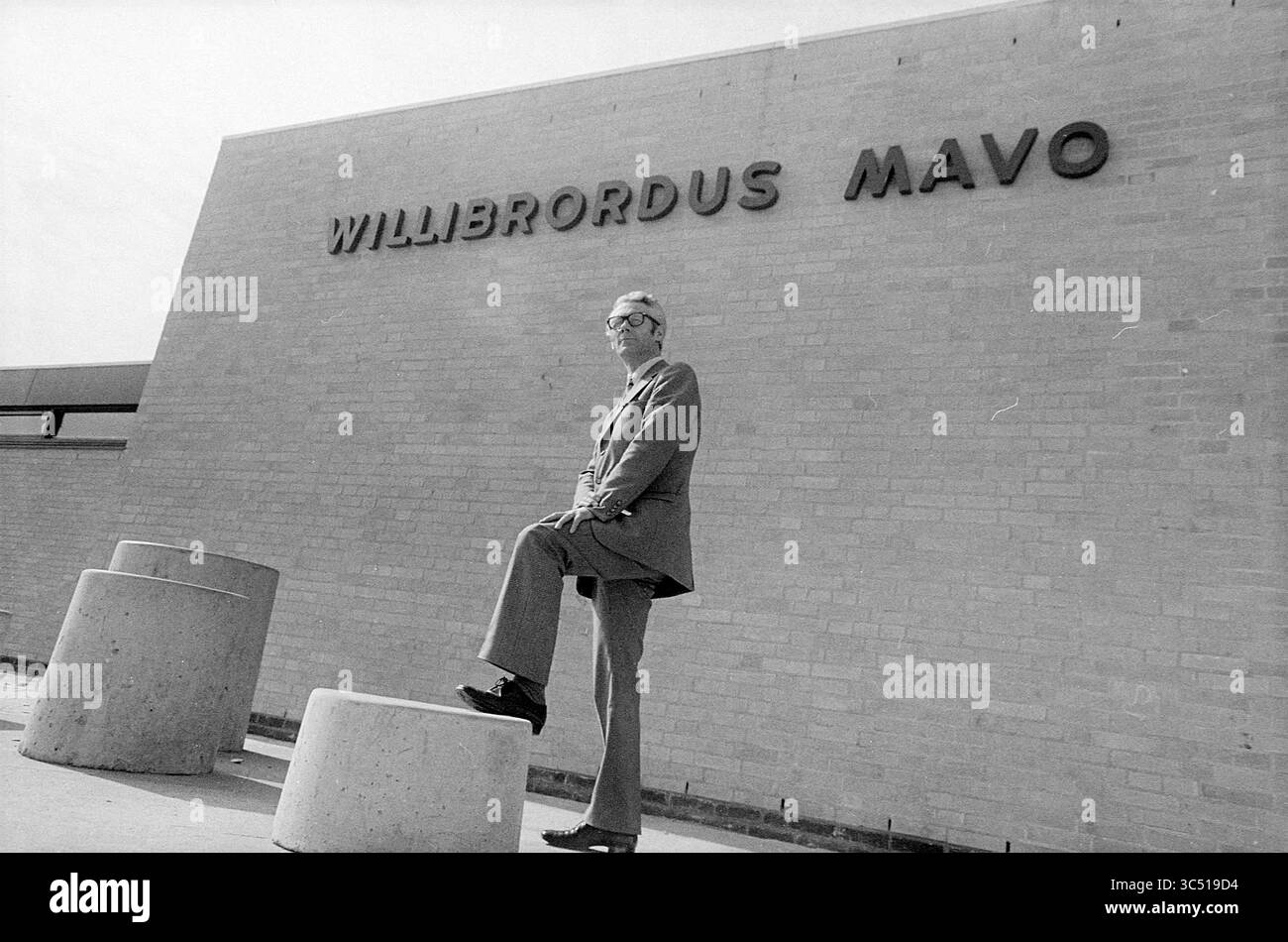 Leiterin Der Williebroius-Schule Mavo Beverwijk, Schulen, 06-04-1973 Whizgle News, Dutch Desk, Niederlande, 1950-2000 Ein Mann im Anzug steht selbstbewusst auf einer Betonbarriere und posiert mit einem Fuß hoch vor einem Gebäude, auf dem der Name „WilliBordus Mavo“ deutlich sichtbar auf seiner Fassade steht. Die Szene fängt eine Mischung aus Stil und architektonischer Einfachheit ein. Stockfoto
