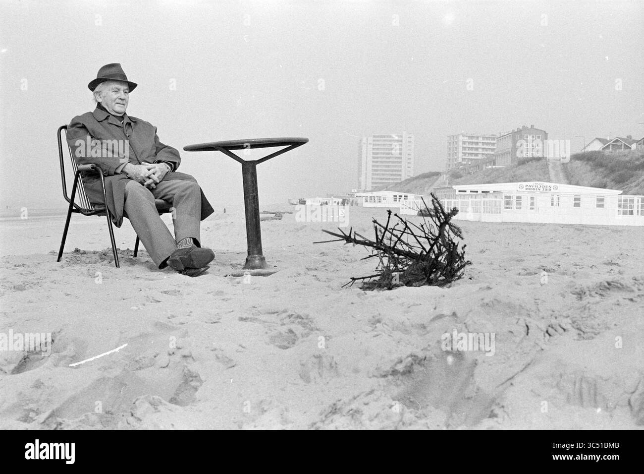 Mann am Strand, Menschen, Strand und Strände, 01-03-1972 Whizgle News, Dutch Desk, Niederlande, 1950-2000 ein älterer Mann sitzt nachdenklich auf einem Stuhl am Strand und blickt auf einen kleinen Treibholzhaufen in der Nähe. Die ferne Küste besteht aus einer Reihe von in Nebel gehüllten Küstengebäuden, die eine ruhige, aber melancholische Atmosphäre schaffen. Stockfoto