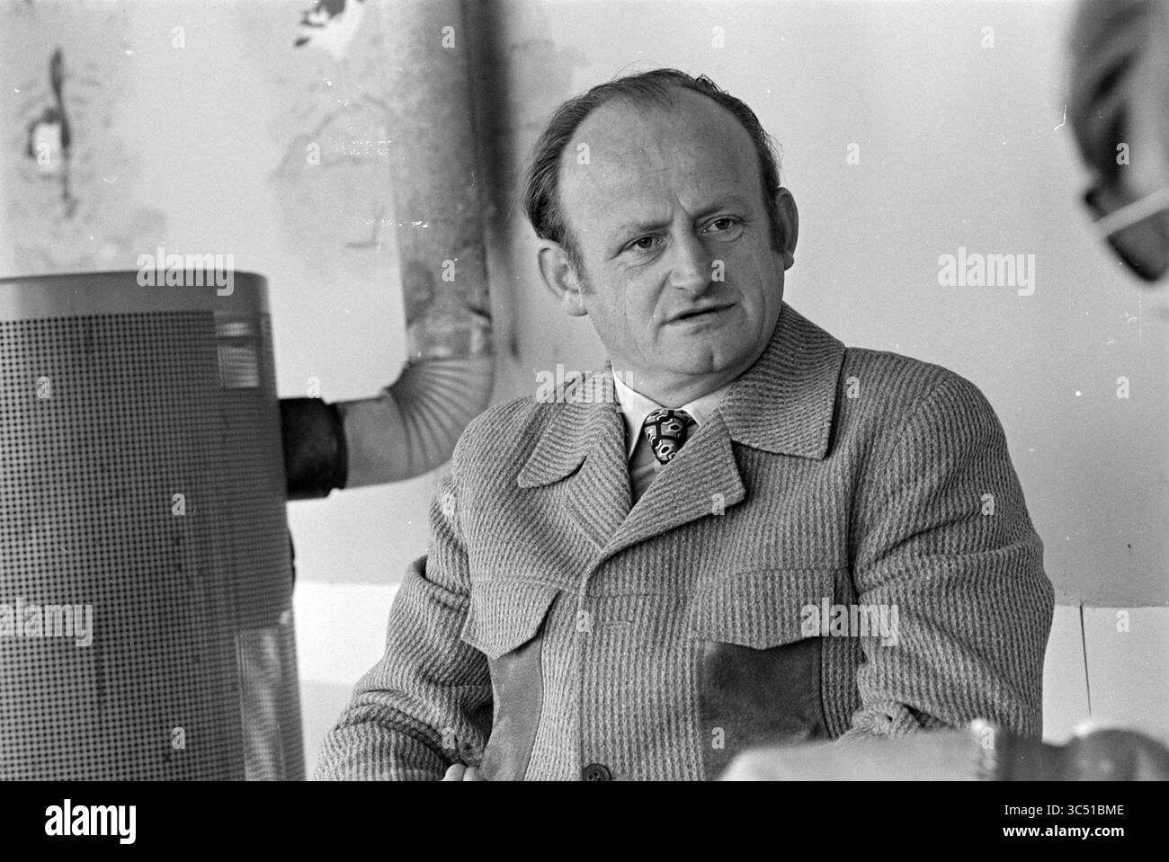 Parnassia mit Mr. Doodemans, Beach and Beaches, 02-10-1978 Whizgle News, Dutch Desk, Niederlande, 1950-2000 Ein Mann in einem maßgeschneiderten Anzug mit einem auffälligen Muster auf seiner Krawatte sitzt und sich in Gesprächen einlässt. Sein Ausdruck ist nachdenklich, während er aufmerksam zuhört, mit einem industriellen Hintergrund, der der Szene Kontext verleiht. Stockfoto